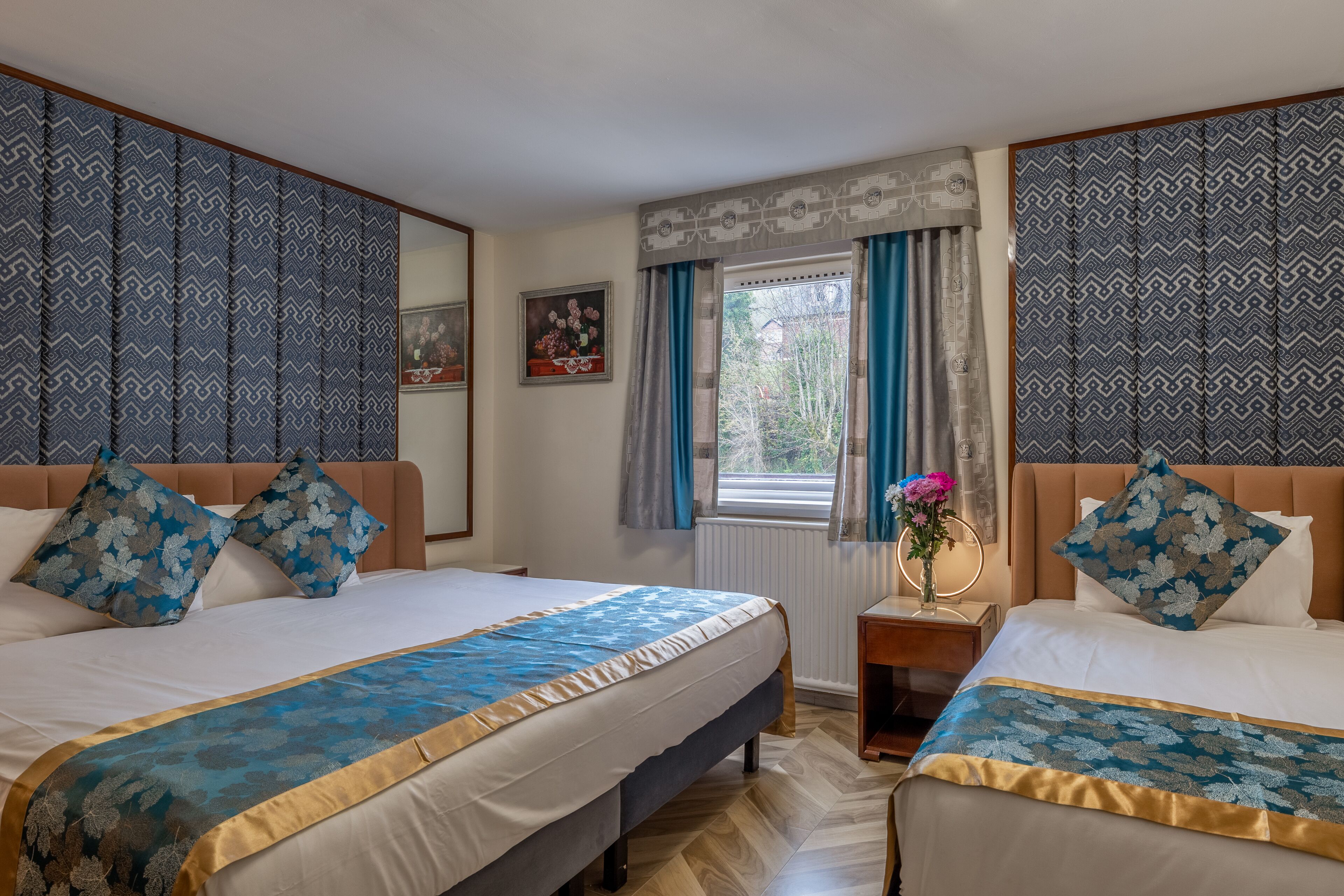 A double room with two beds, each with a patterned bedspread and matching pillows. There is a window with curtains, a bedside table with a vase of flowers, and a framed picture on the wall.