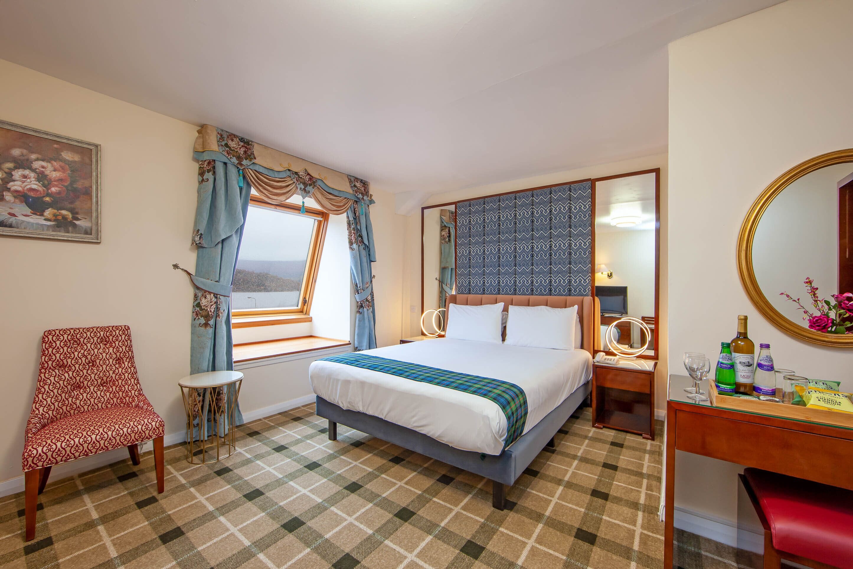 A hotel room with a bed, a chair, a small table, a mirror, and a window with curtains.