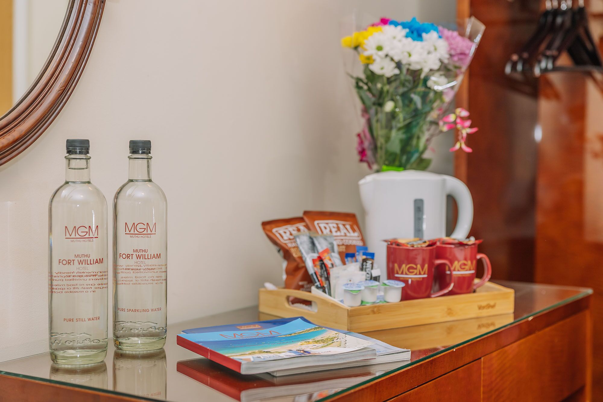 A hotel reception area with two bottles of water, a coffee maker, a tray with coffee packets, a vase with flowers, and a magazine on the counter.