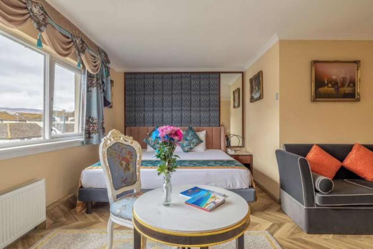 A hotel room with a bed, a sofa, a round table with a vase and book, and a window with curtains.