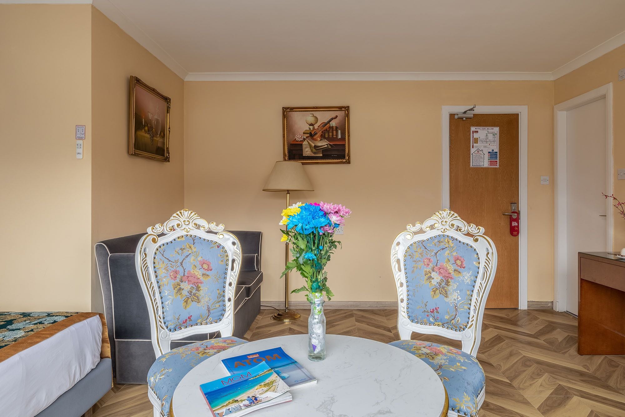 A hotel room with two ornate chairs, a small round table with a vase of flowers, a bed with a blue floral bedspread, and a door with a fire alarm.