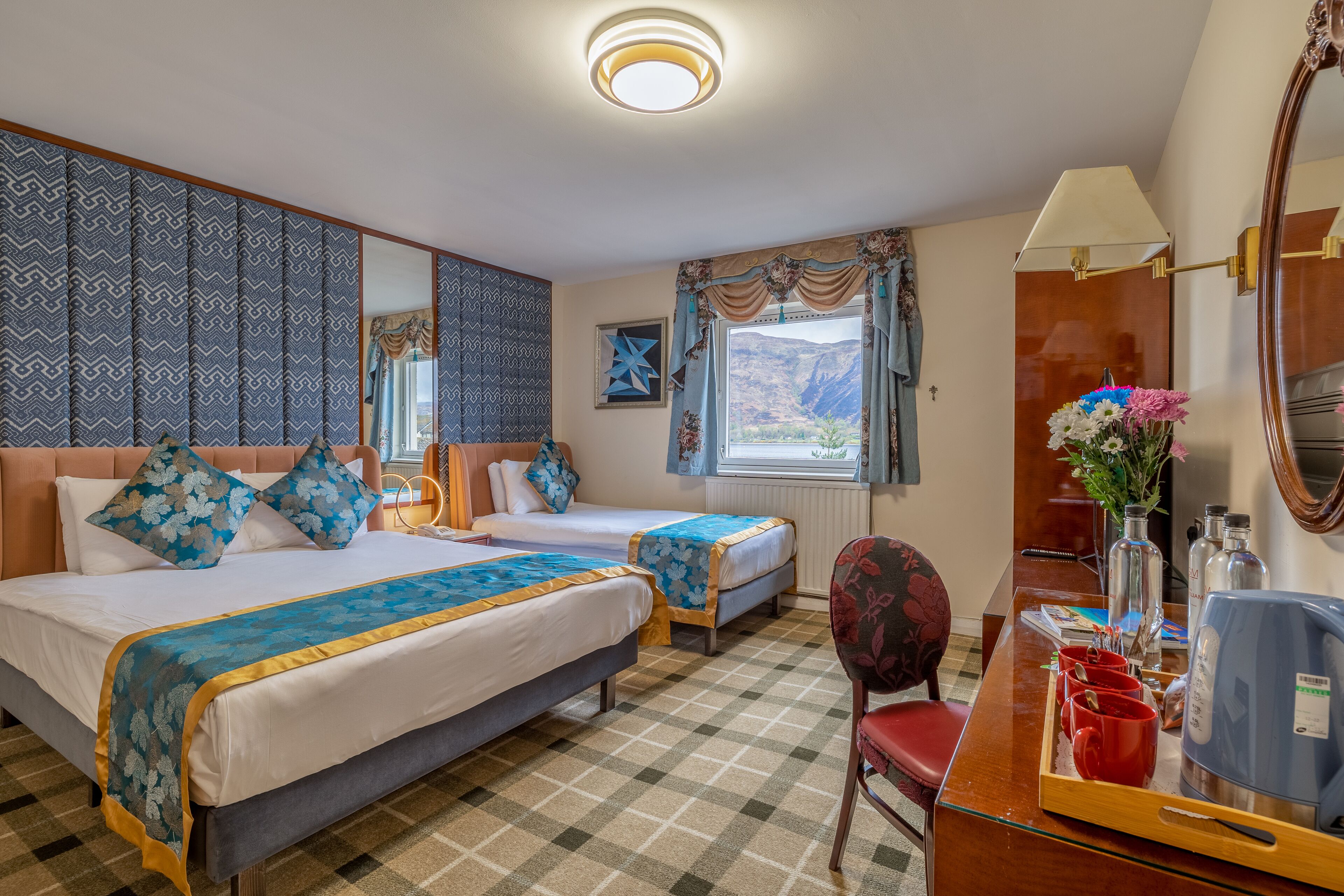 Habitación de hotel con dos camas, un escritorio, una silla, un espejo, una ventana con cortinas y vistas a las montañas.
