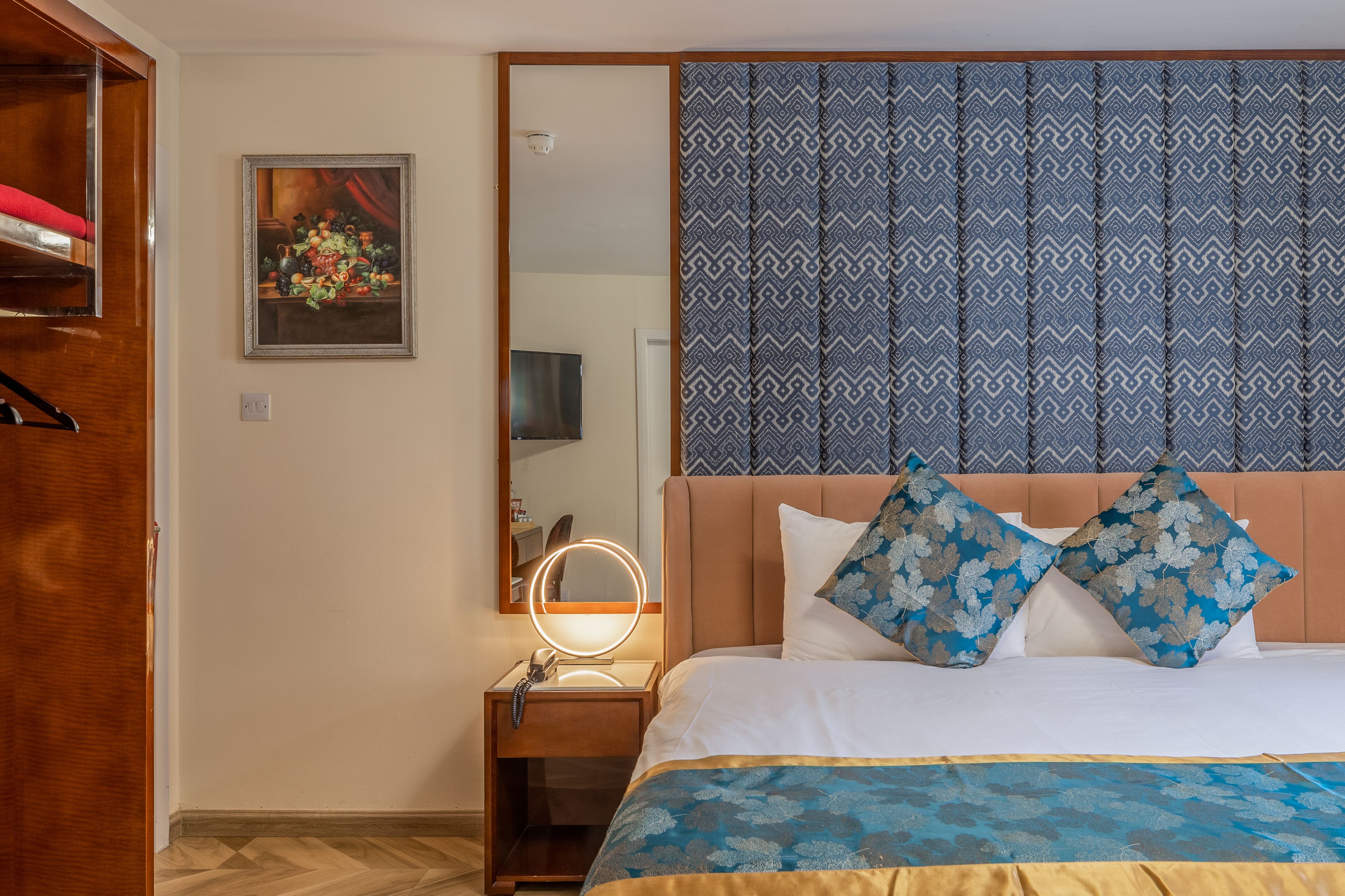 A hotel room with a bed, a nightstand with a mirror, a patterned headboard, and a framed painting on the wall.