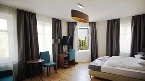 A hotel room with a bed, a desk, a chair, a TV, and large windows with curtains. - Bett & Buch Hotel (Berlin)