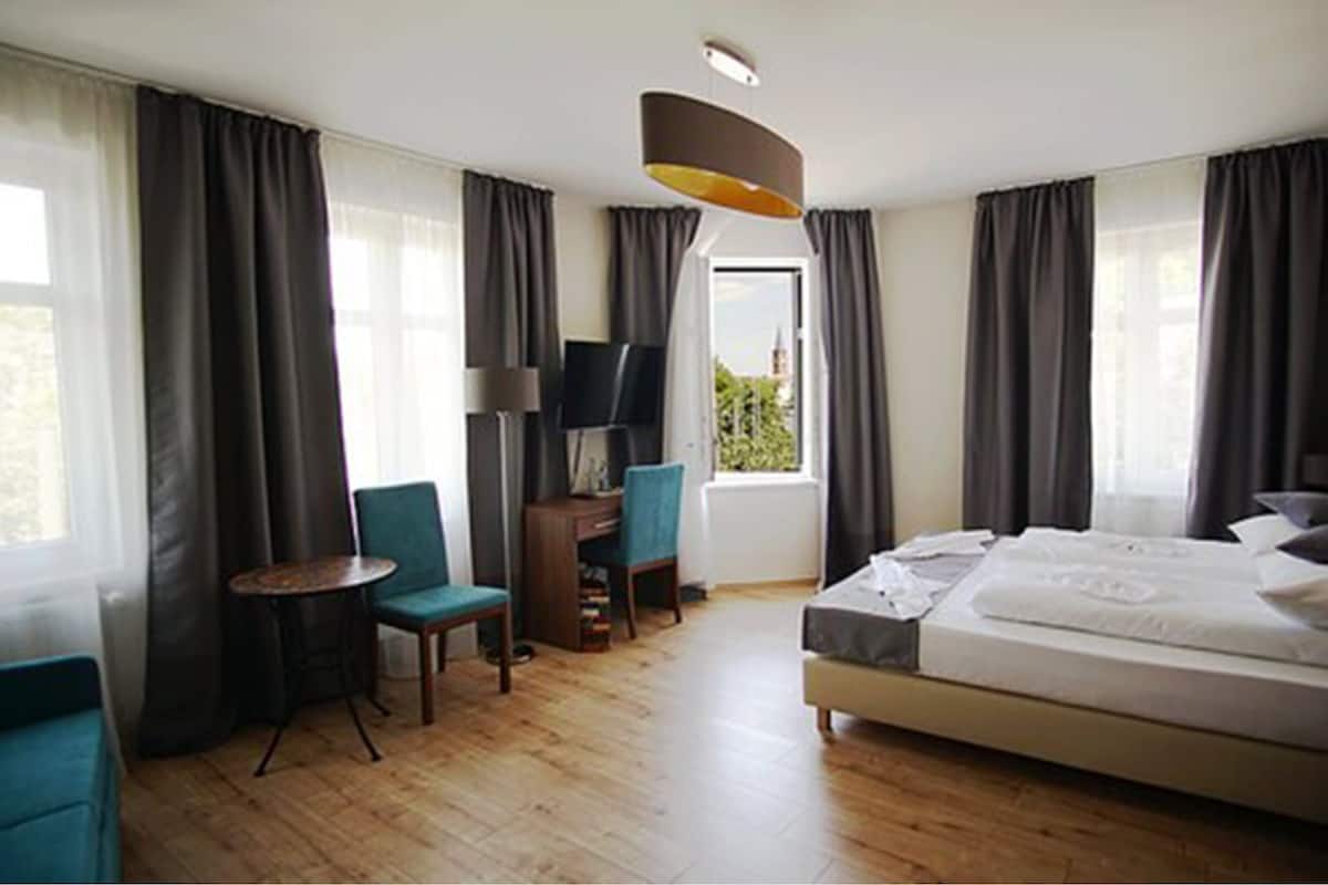 A hotel room with a bed, a desk, a chair, a TV, and large windows with curtains.