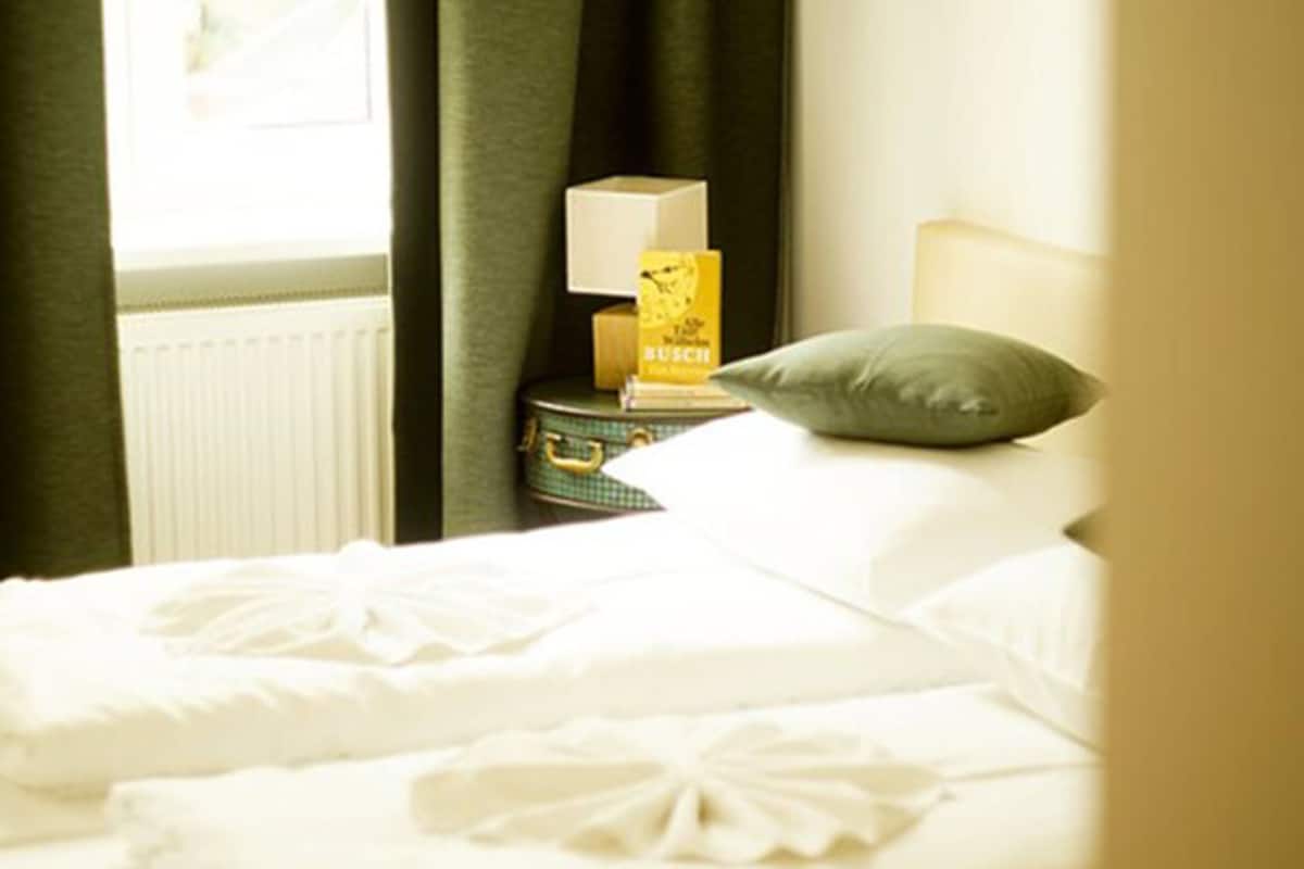A neatly made bed with white linens and a pillow, a bedside table with a lamp and a book, and a window with curtains.