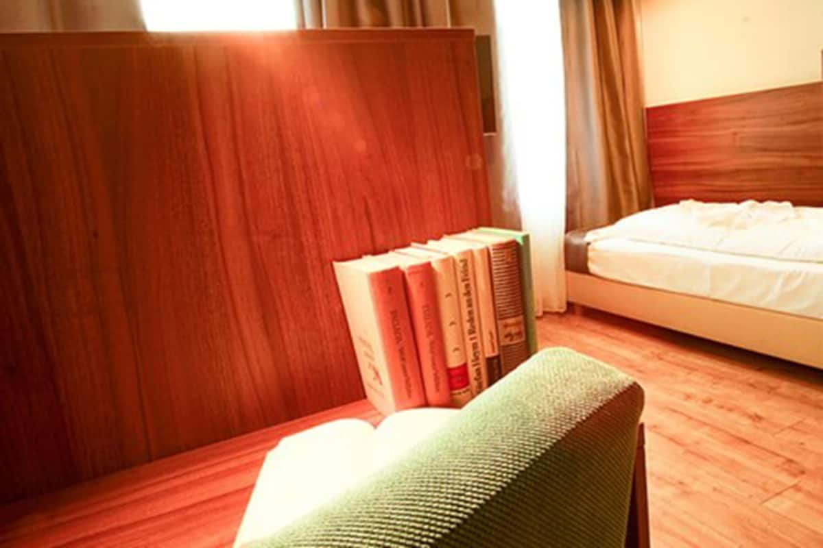 A hotel room with a wooden headboard, a bed with white bedding, and a green chair with books on a wooden table.