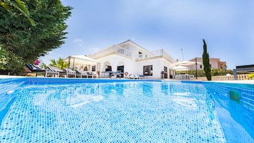 Une piscine avec une terrasse carrelée, des chaises longues et un bâtiment à l’arrière-plan, doté d’une façade blanche et de balcons.