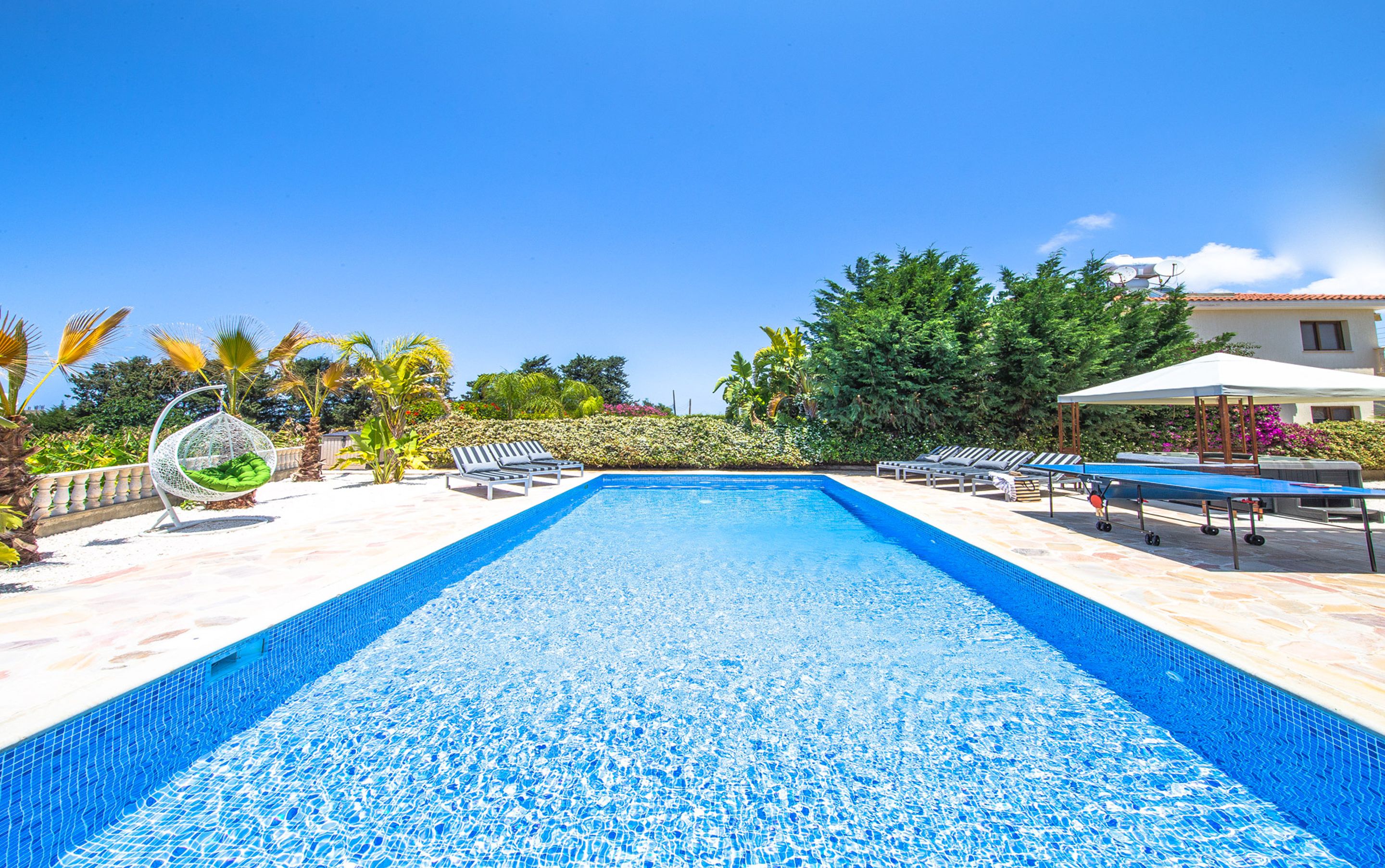 Una piscina con lettini e una sedia a dondolo, immersa nel verde sotto un cielo sereno.