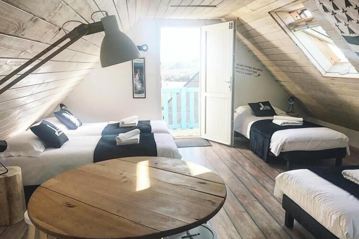 Ein gemütliches Schlafzimmer im Dachgeschoss mit zwei Betten, einem Holztisch und einem großen Fenster mit Blick ins Freie.