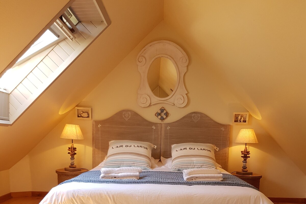 Ein ordentlich bezogenes Bett mit Kissen, beschriftet mit "L'Air du Large", in einem Zimmer mit Holzboden, geneigter Decke und zwei Nachttischlampen.
