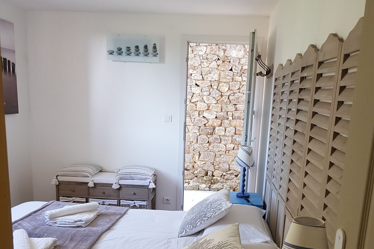Ein Schlafzimmer mit einem Bett, einem Nachttisch, einer Lampe, einem Fenster mit Blick auf eine Steinmauer und einer Wanddekoration.