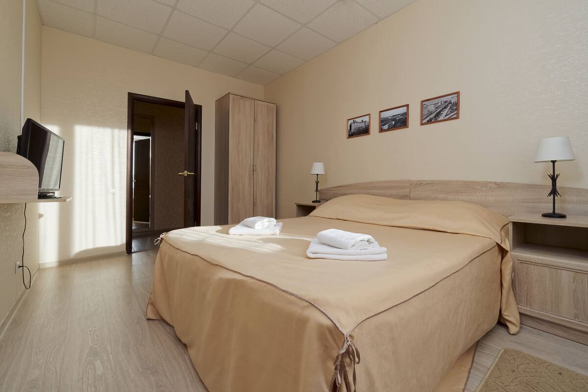A hotel room with a bed, a wardrobe, a desk with a lamp, and three framed pictures on the wall.