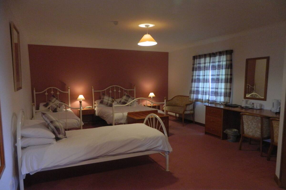 A hotel room with two single beds, a desk with a chair, a small table, and a window with curtains.