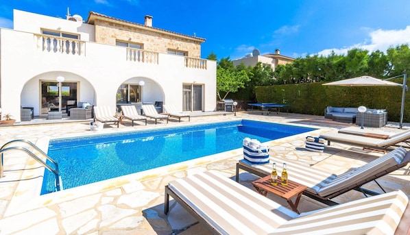 A pool area with lounge chairs, a barbecue, and a white building with arches.