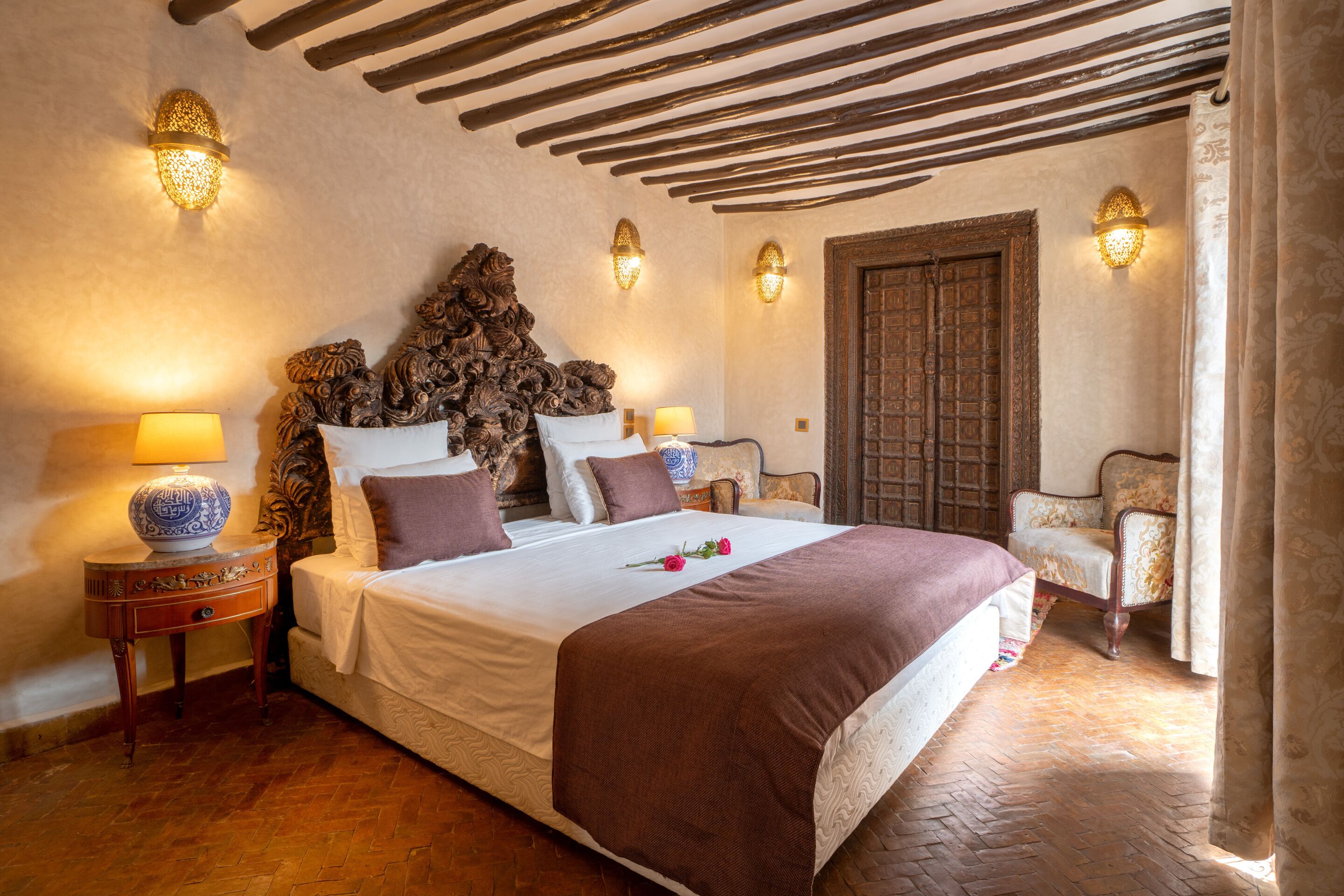 Un dormitorio con una cama grande, cabecera de madera, apliques decorativos en la pared, una puerta de madera y una silla.