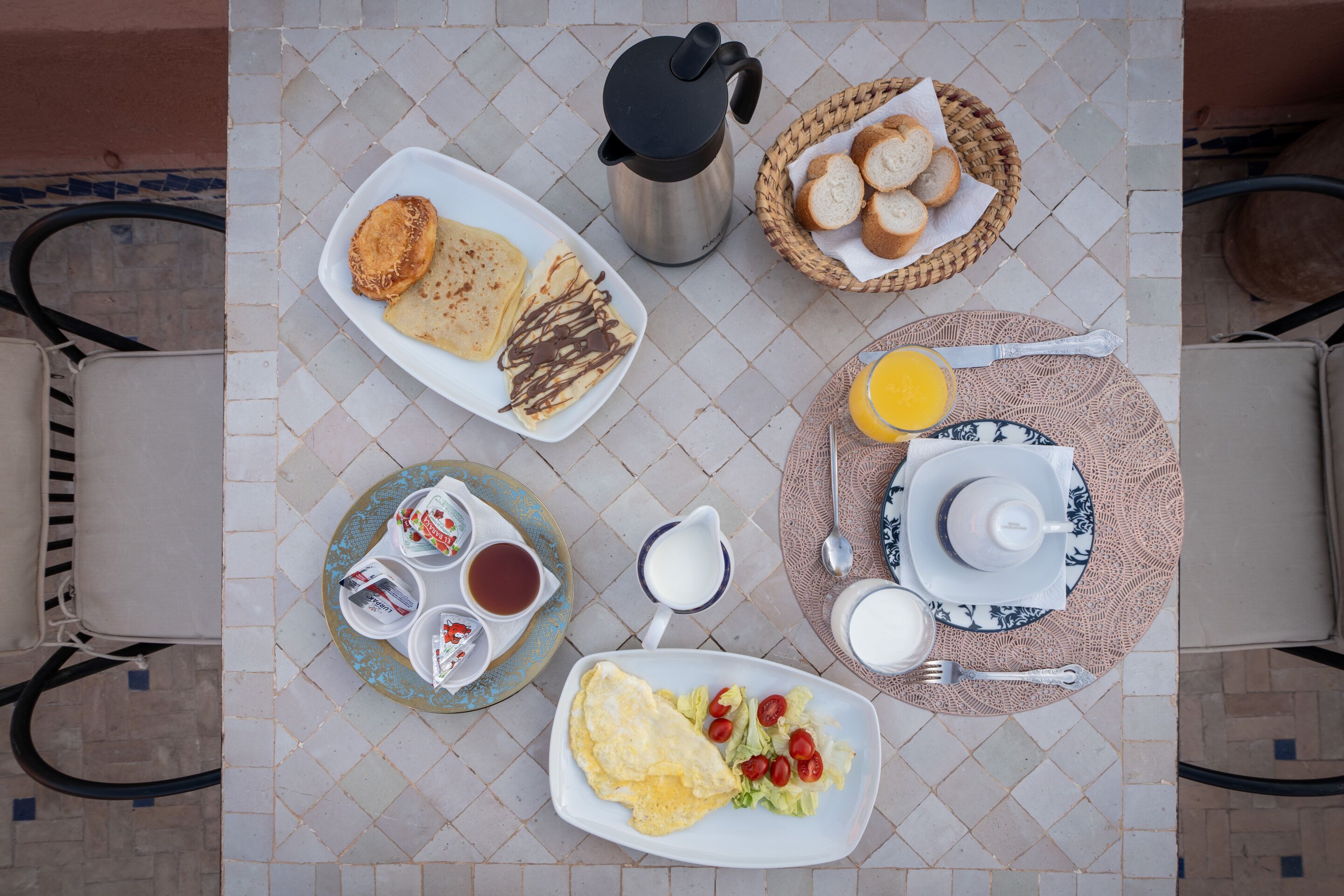 Una mesa de desayuno con diversos platos, como panqueques, tostadas, huevos y fruta. Hay una cafetera, una cesta de pan y un plato con jugo de naranja.