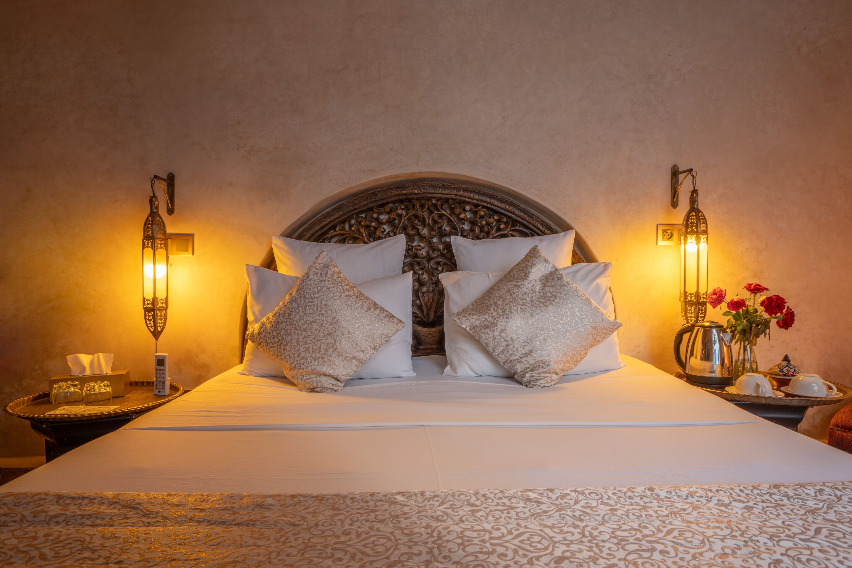 Una cama bien hecha con almohadillas estampadas y un cabecero decorativo, una mesita de noche con una caja de pañuelos y un florero, y lámparas fijadas a la pared.