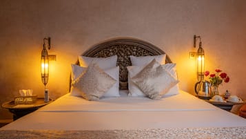 Una cama bien hecha con almohadillas estampadas y un cabecero decorativo, una mesita de noche con una caja de pañuelos y un florero, y lámparas fijadas a la pared.