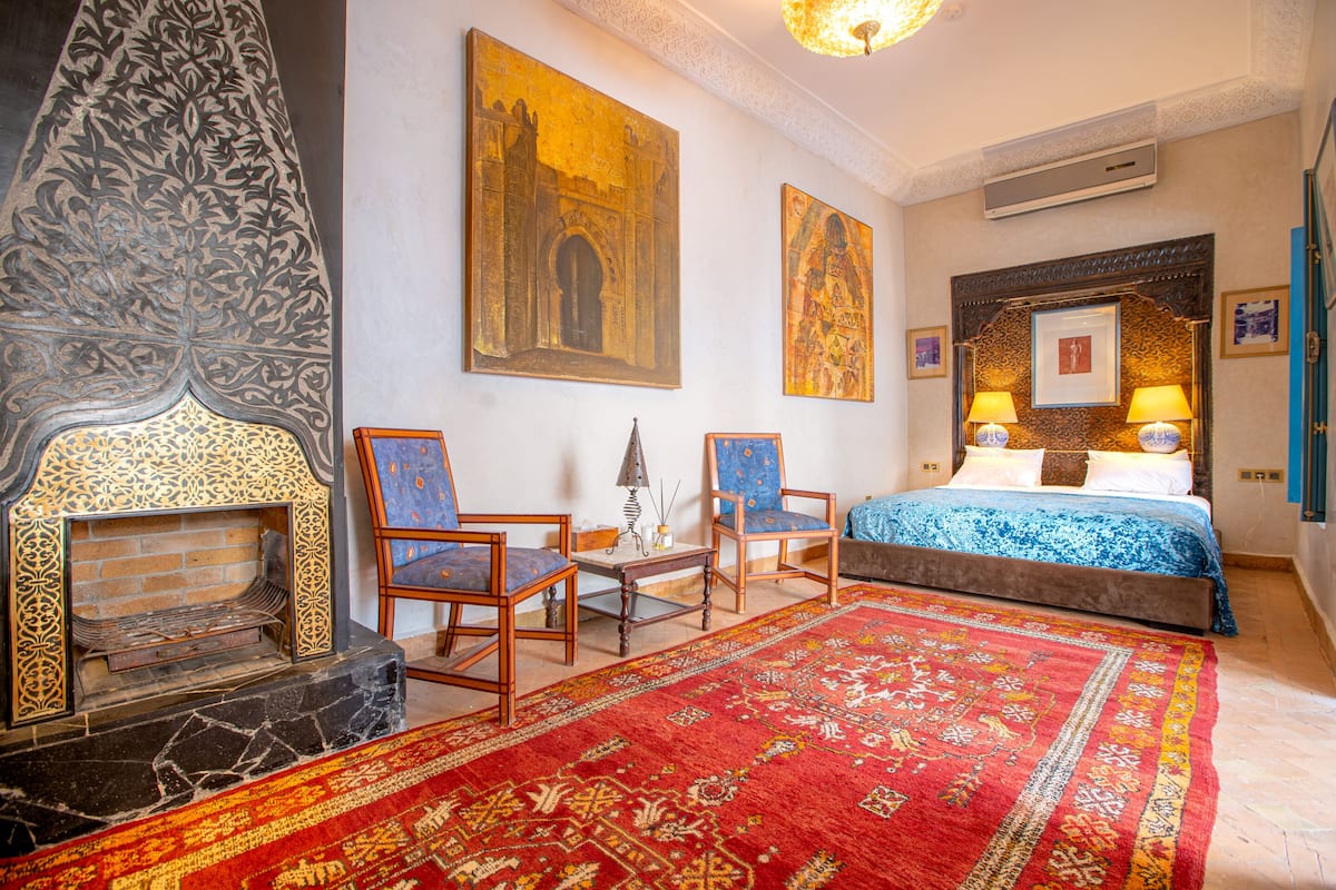 Een kamer met een open haard, twee stoelen, een bed met een blauwe sprei en een rode tapijt.
