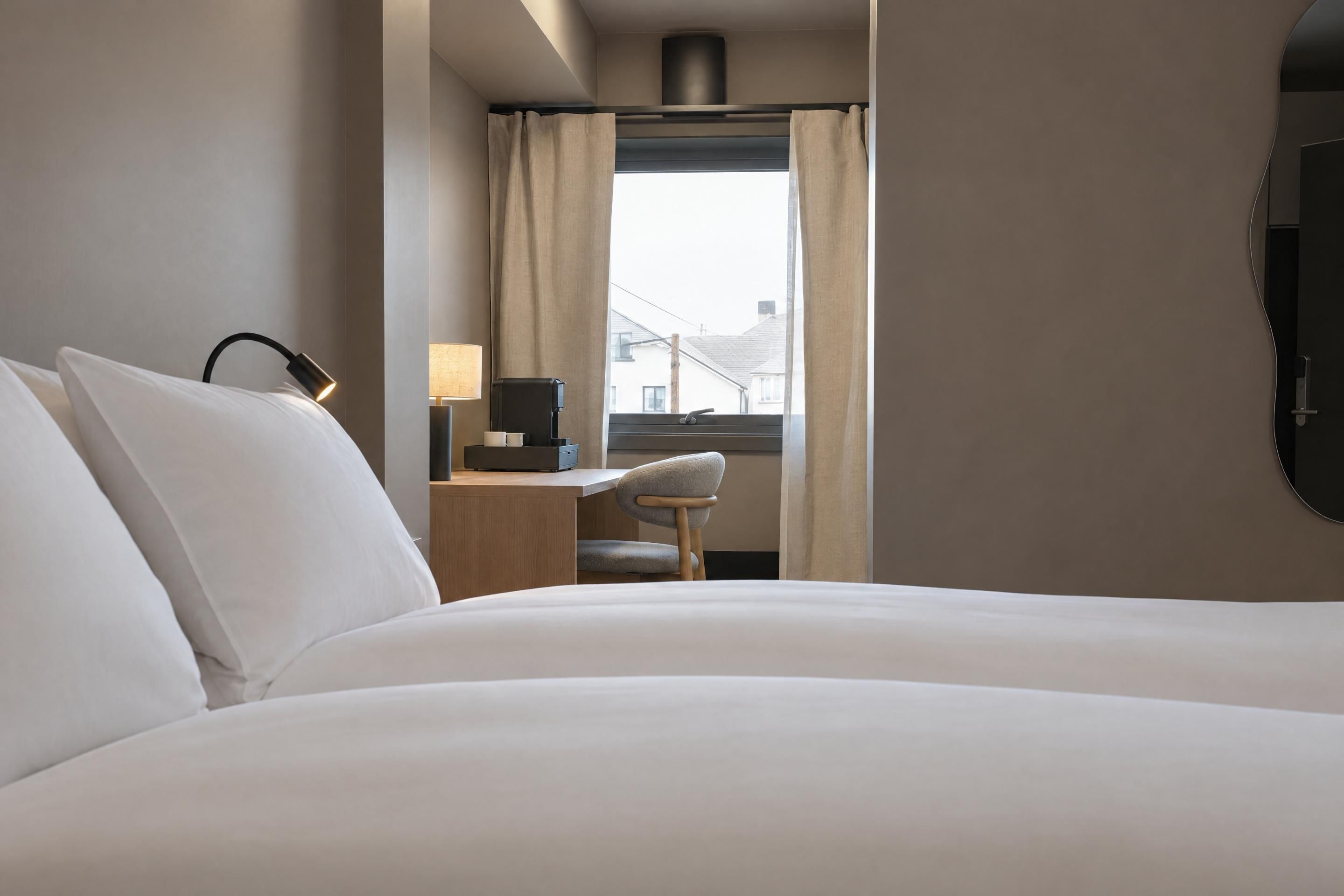 A hotel room with a bed, a desk with a lamp, a chair, and a window with curtains.