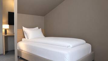 Una cama bien tendida con ropa blanca en una habitación con techo inclinado y una mesita de noche con una lámpara.