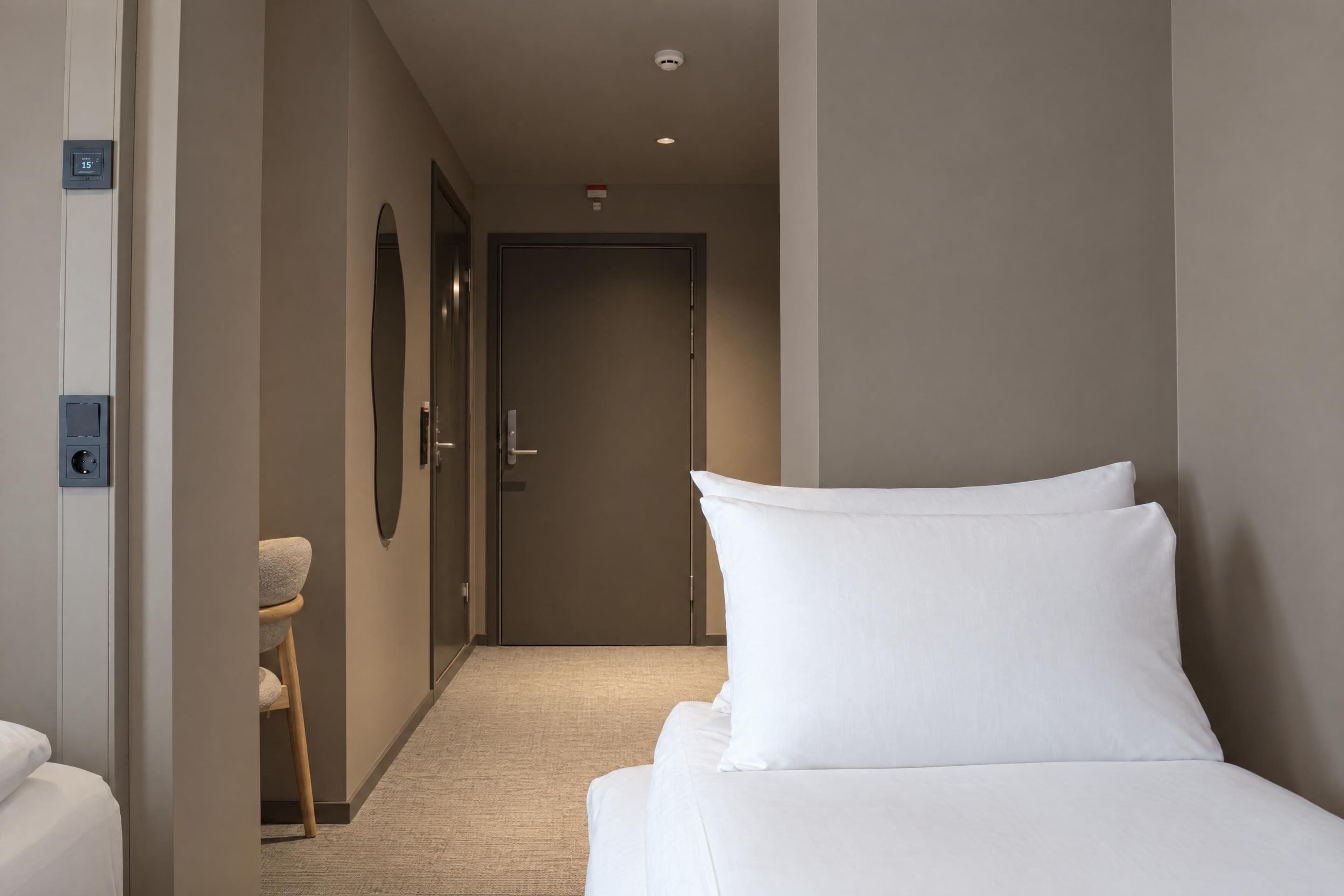 Habitación de hotel con cama, una silla, una puerta y un termostato en la pared.