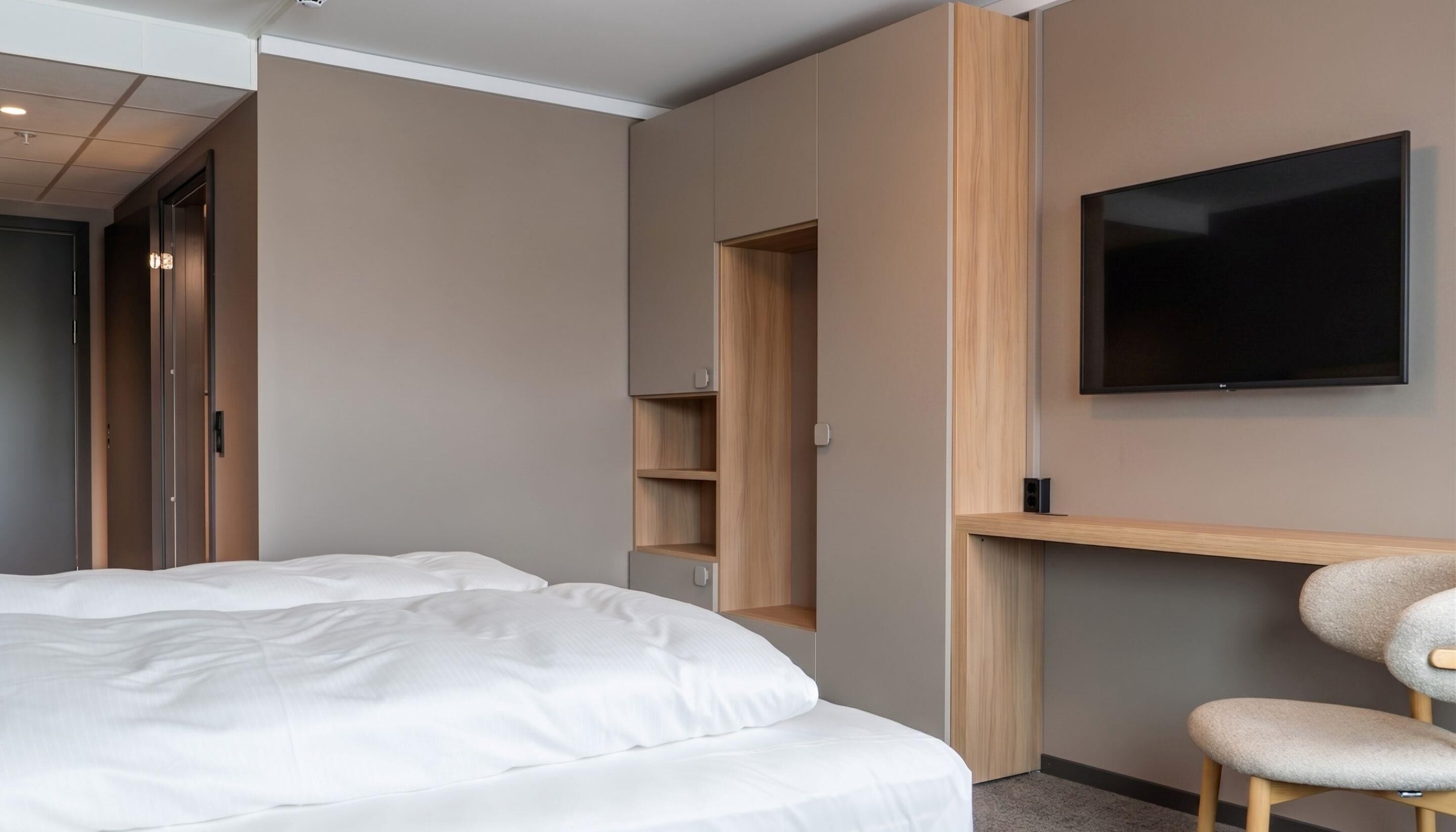 Una habitación de hotel con una cama, un televisor montado en la pared y una estantería de madera.