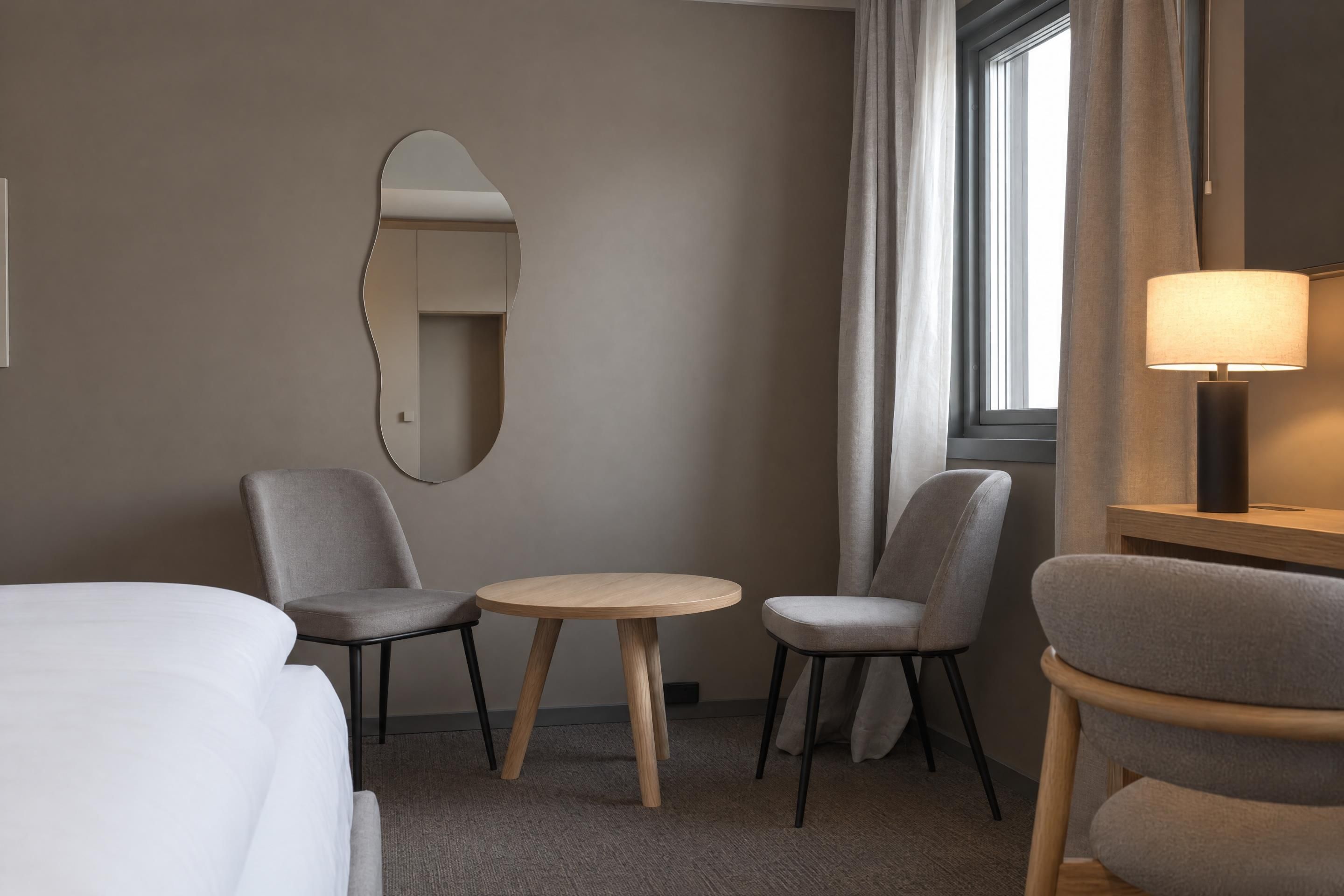 A hotel room with a bed, two chairs, a small round table, a mirror, a lamp, and a window with curtains.