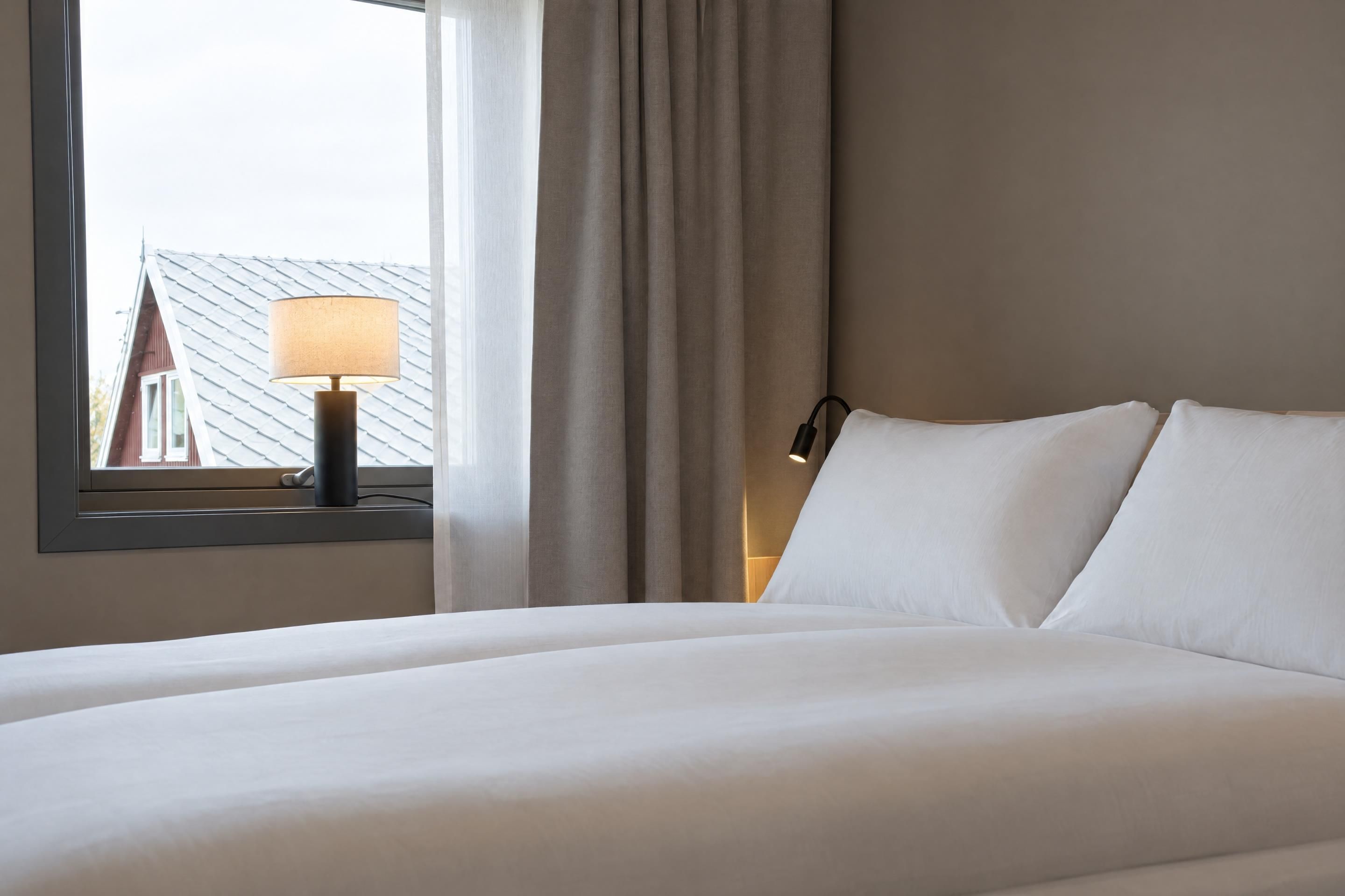 Een hotelkamer met een bed, een raam met gordijnen, een lamp en uitzicht op een gebouw.