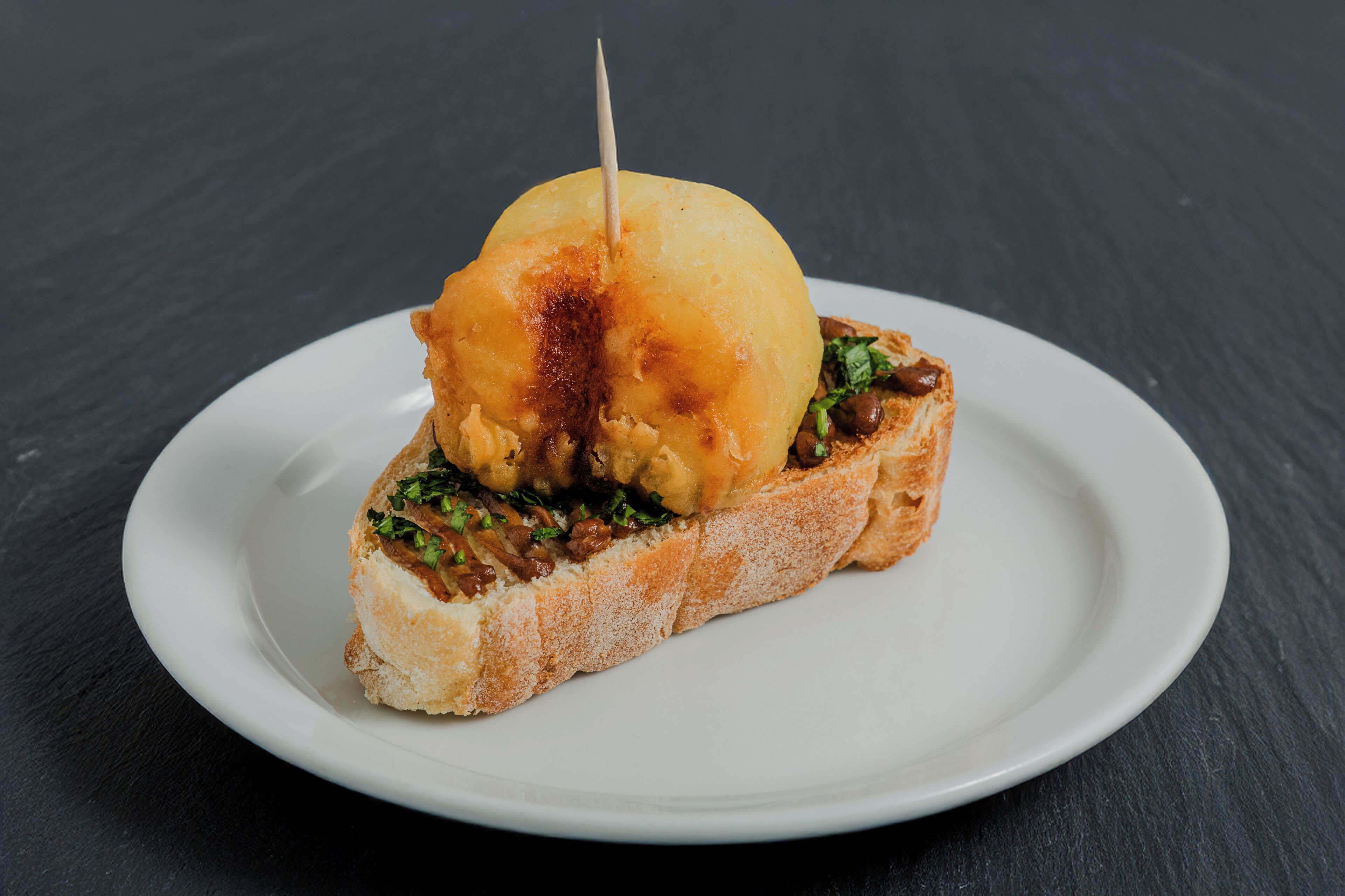 A sandwich on a white plate with a toothpick, placed on a dark surface.