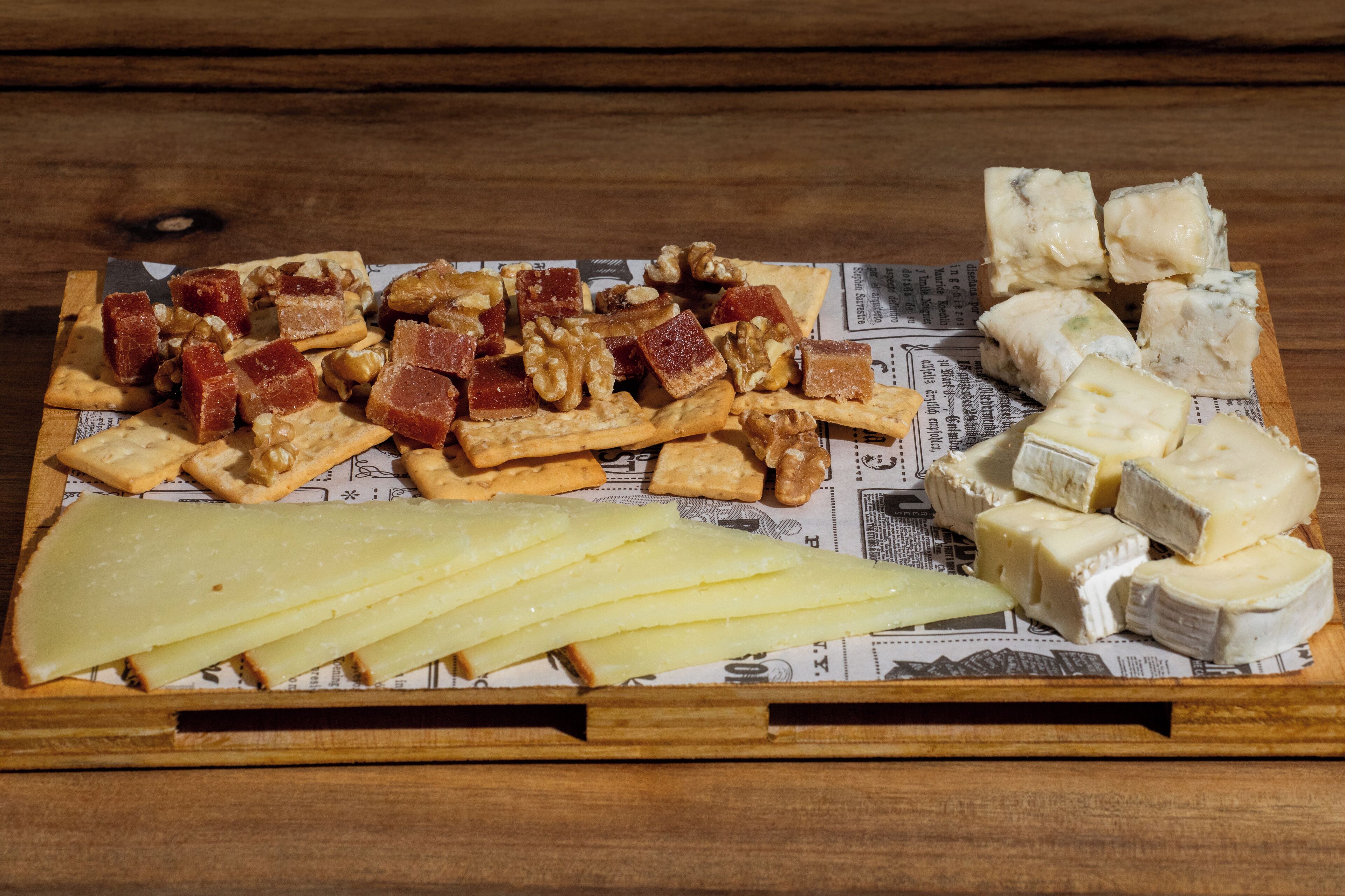 A wooden tray with various cheeses, crackers, and nuts on a newspaper background.