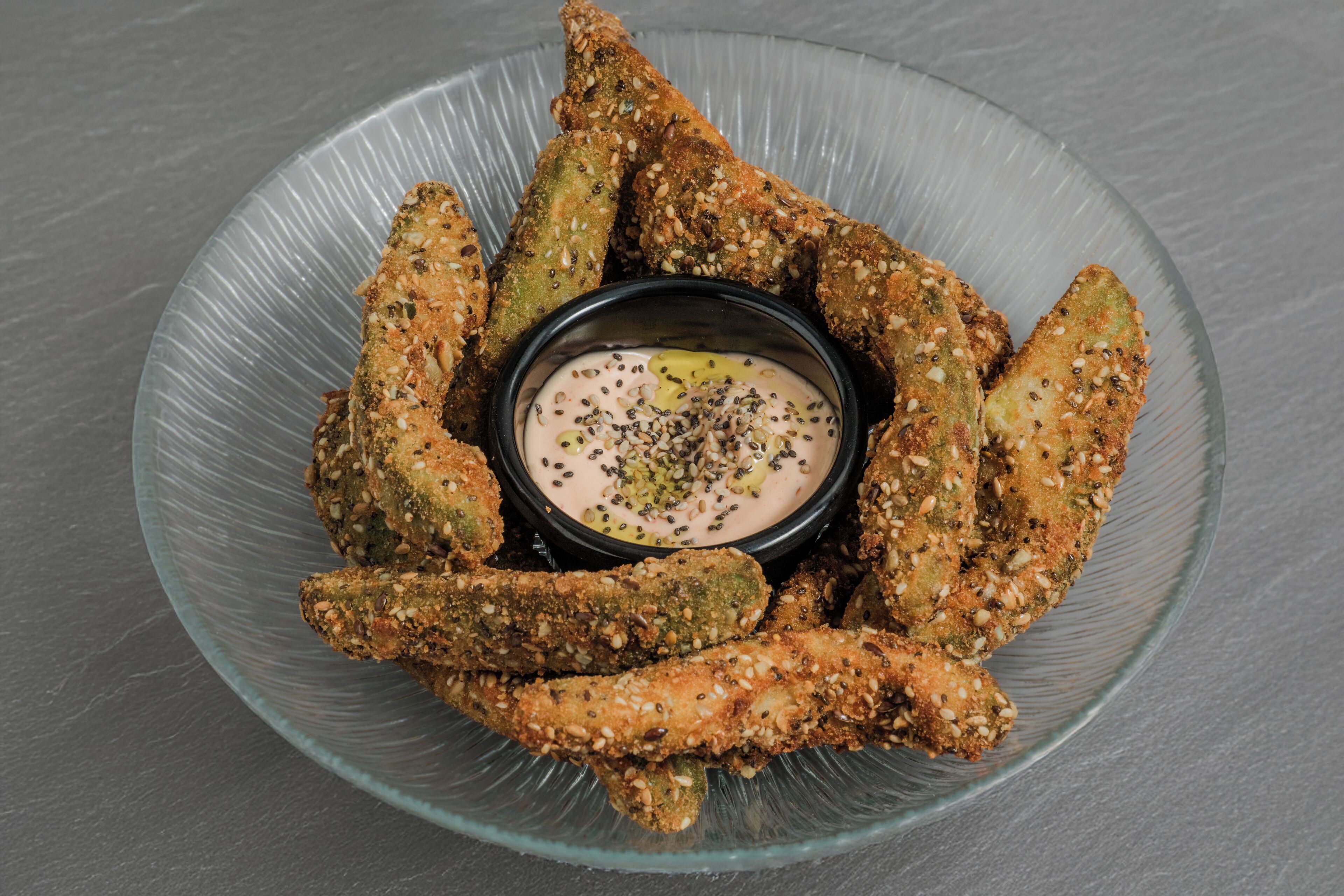 Un bol en verre contenant des bâtonnets de courgettes frits, accompagnés d’une sauce pour tremper, le tout entouré d’autres bâtonnets de courgettes frits sur une surface grise.