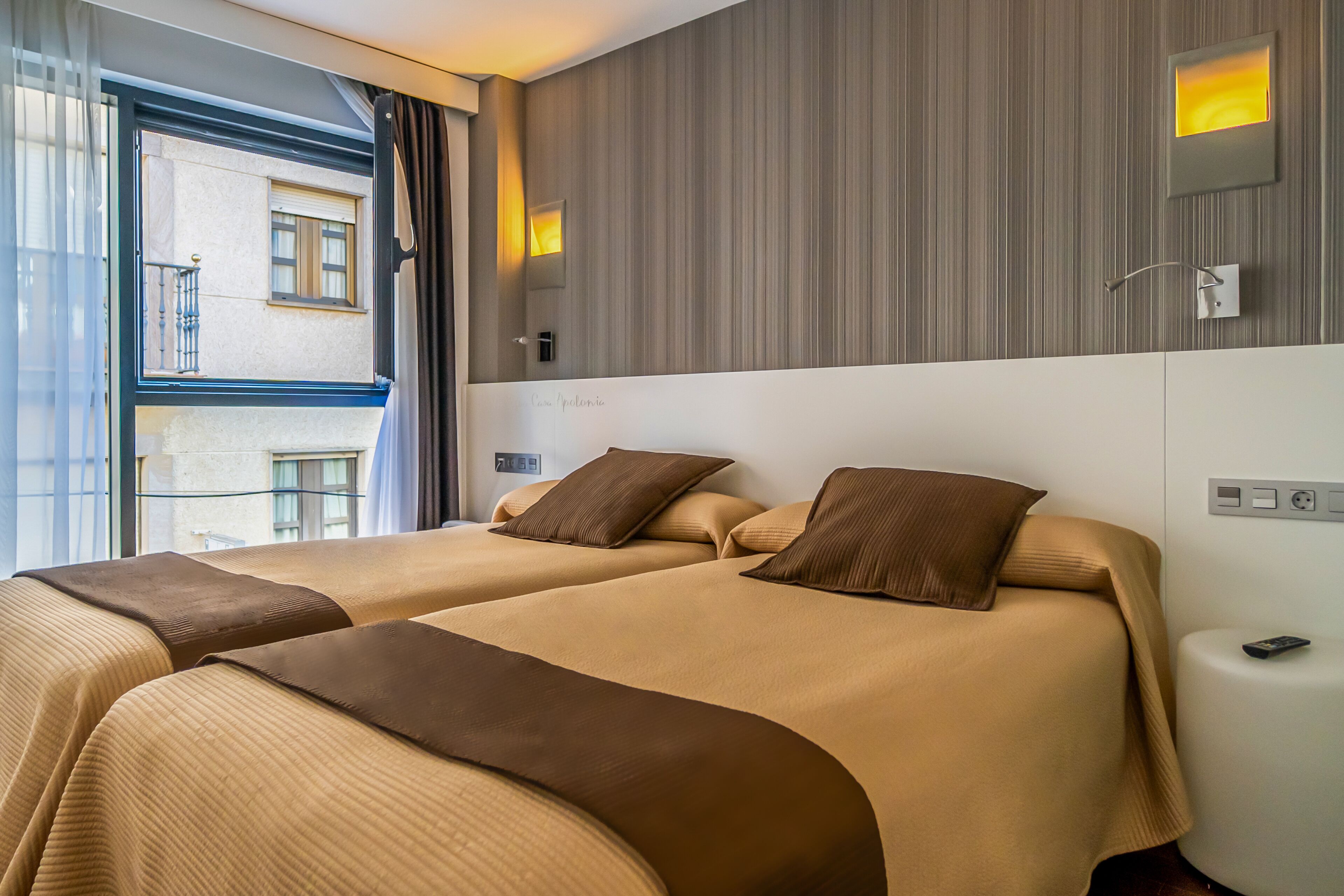 A hotel room with two beds, a large window, and a wall-mounted light fixture.