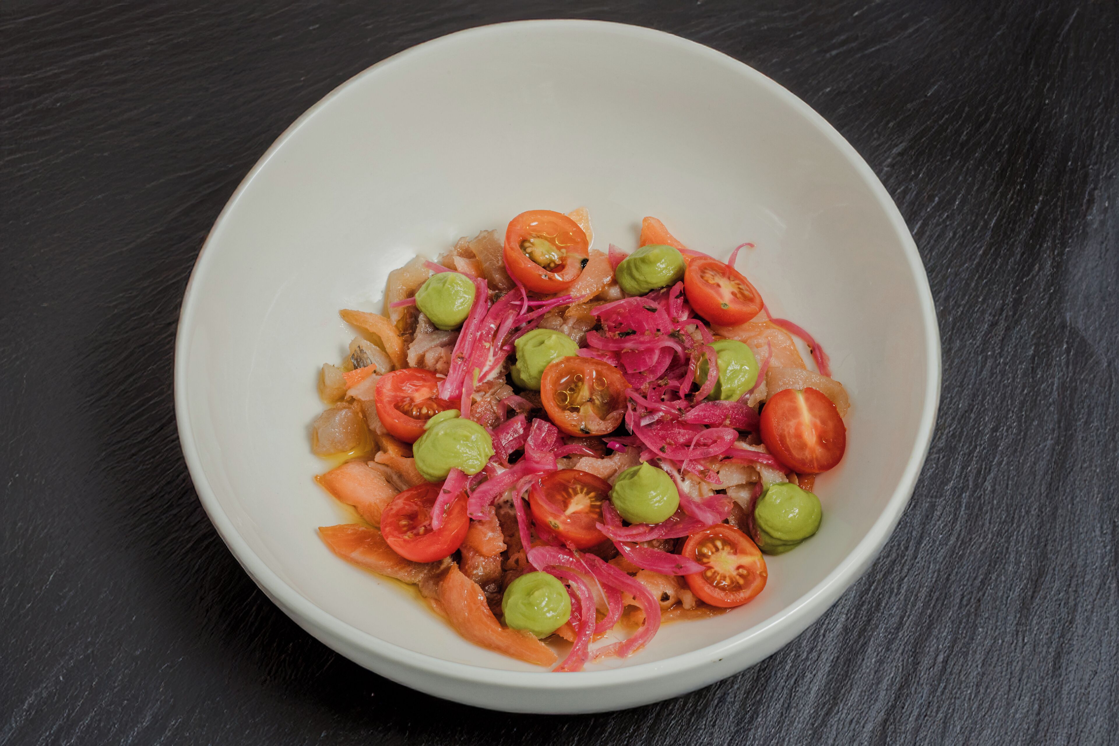 A white bowl containing a salad with tomatoes, onions, and green olives on a dark surface.