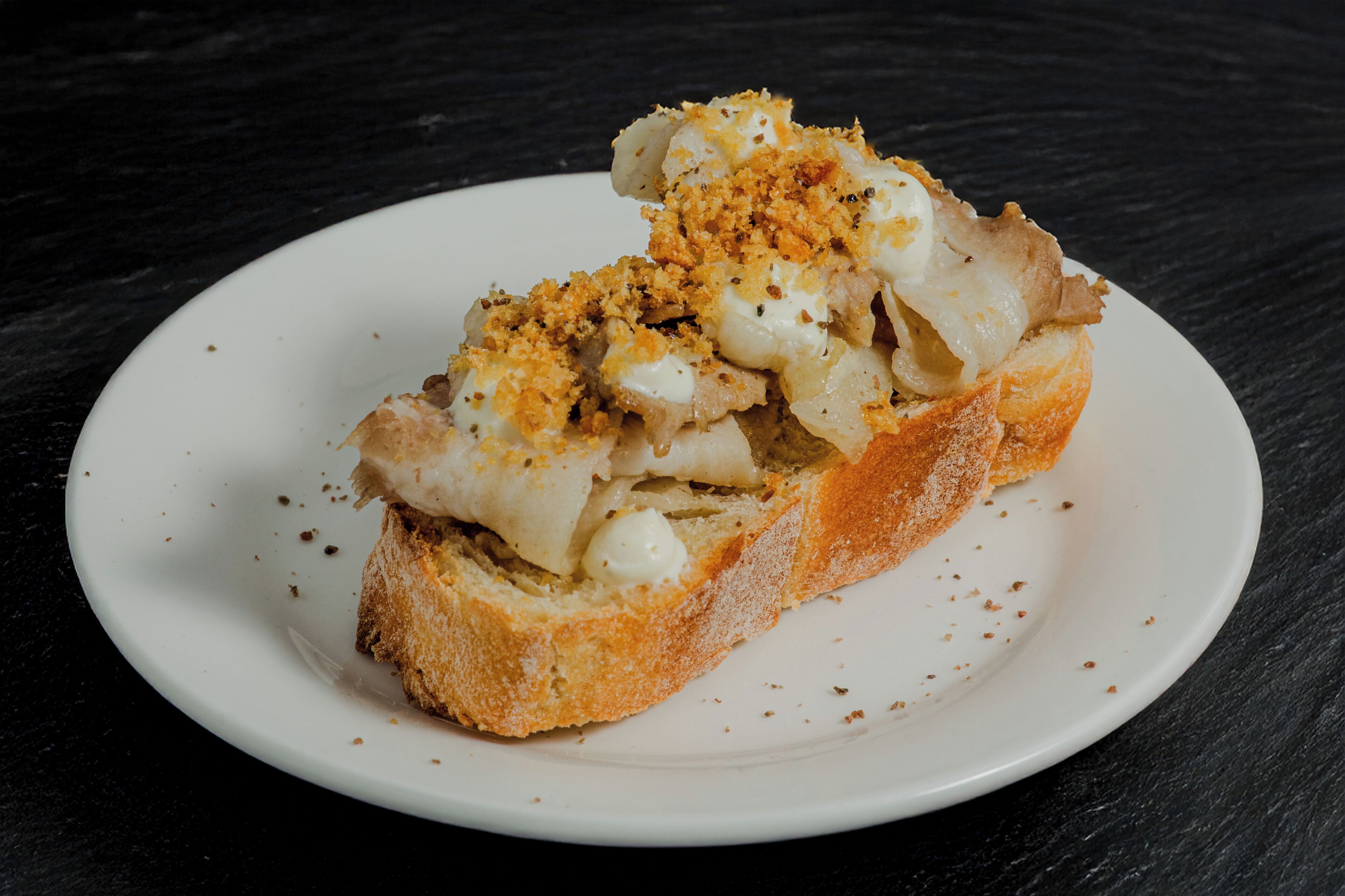 Un plato blanco con una rebanada de pan cubierta con salsa blanca y migas por encima, sobre una superficie oscura.