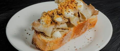 Un plato blanco con una rebanada de pan cubierta con salsa blanca y migas por encima, sobre una superficie oscura.