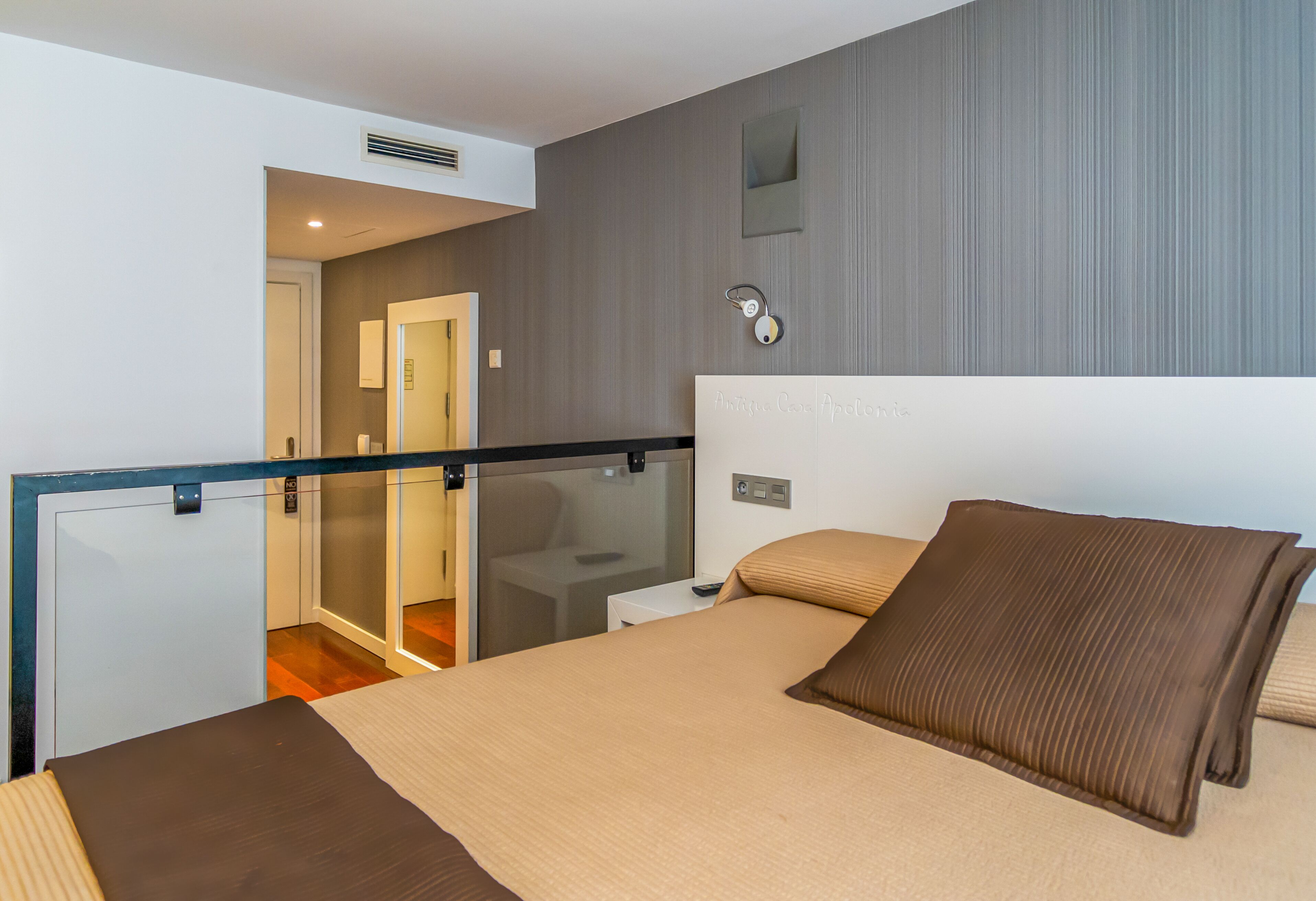 Una habitación de hotel moderna con una cama grande, un separador de vidrio, una mesita de noche pequeña y un aplique de pared.