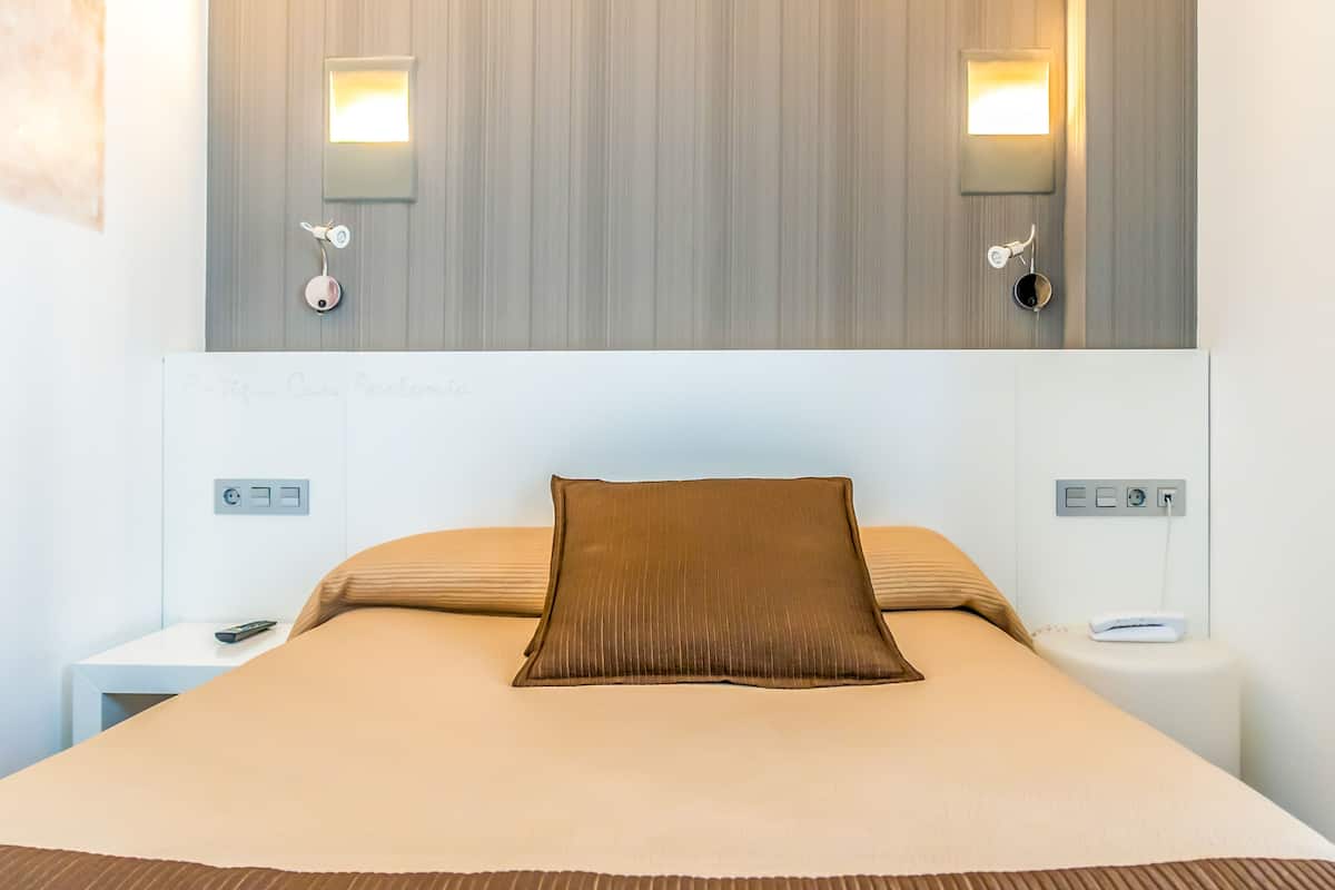 A modern hotel room with a large bed, two bedside tables, a nightstand, and a wall-mounted light fixture.