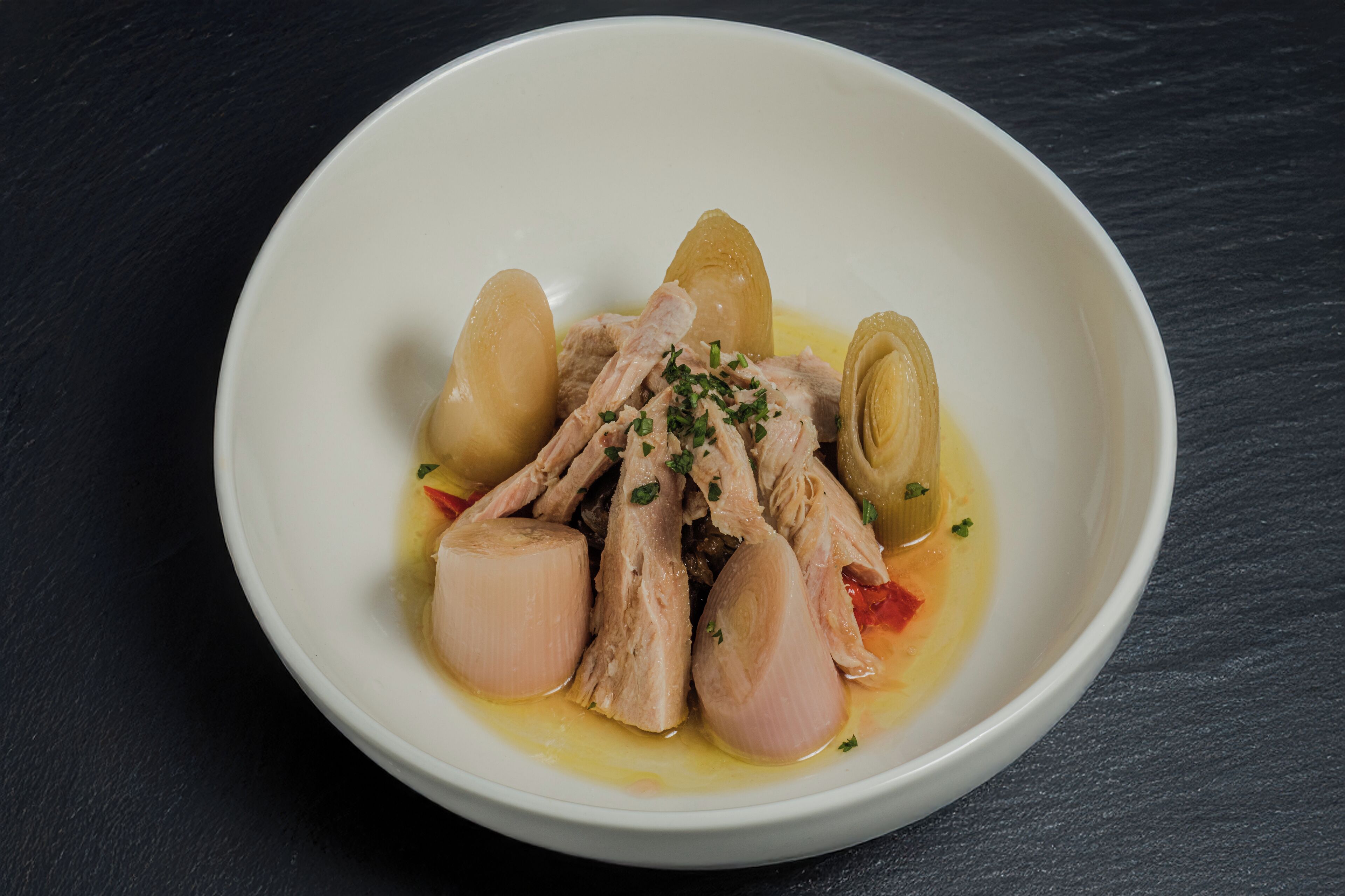 A white bowl containing shredded chicken, potatoes, and a yellow sauce, garnished with herbs, on a dark surface.