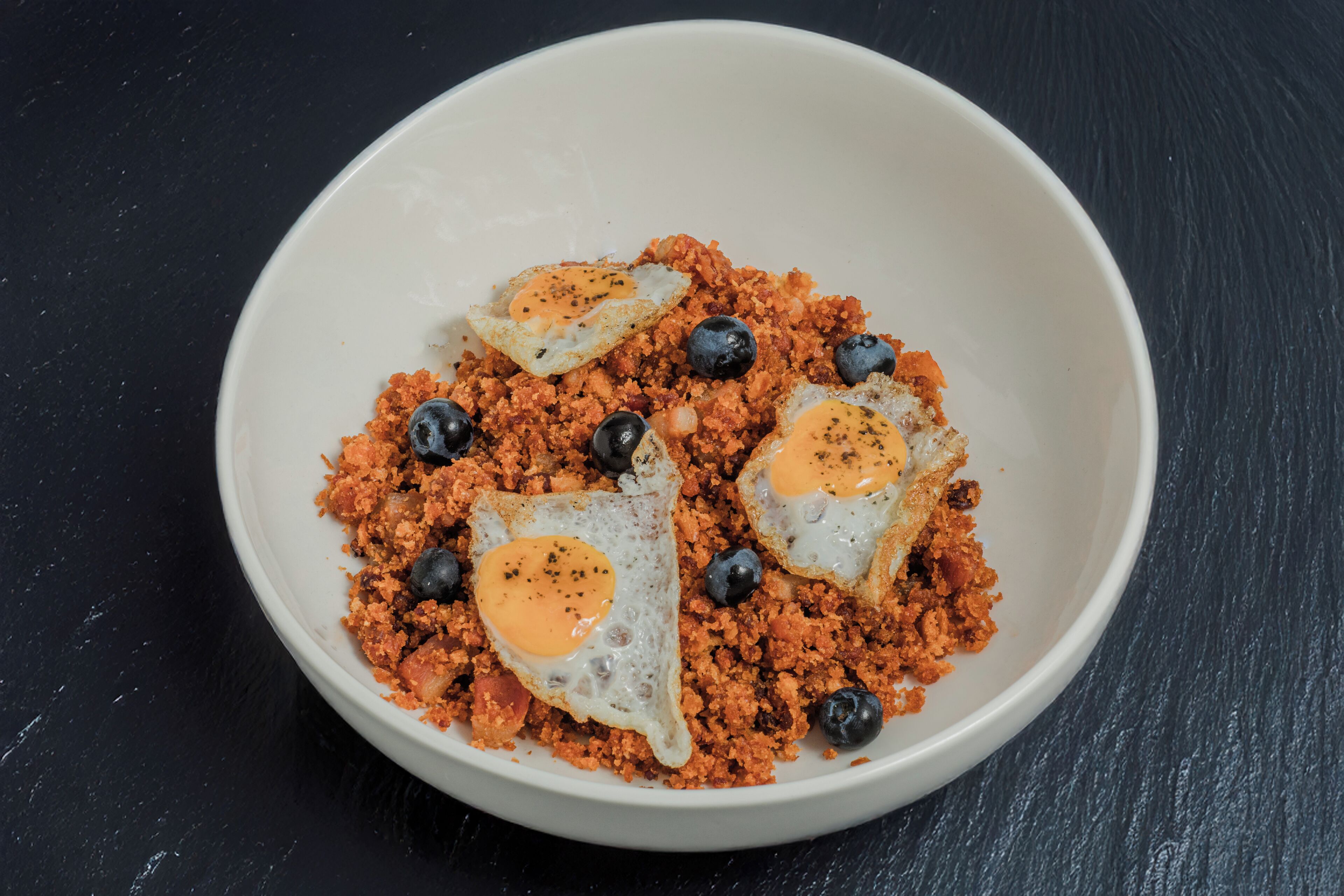 A white bowl containing a dish with ground meat, eggs, and blueberries on a dark surface.