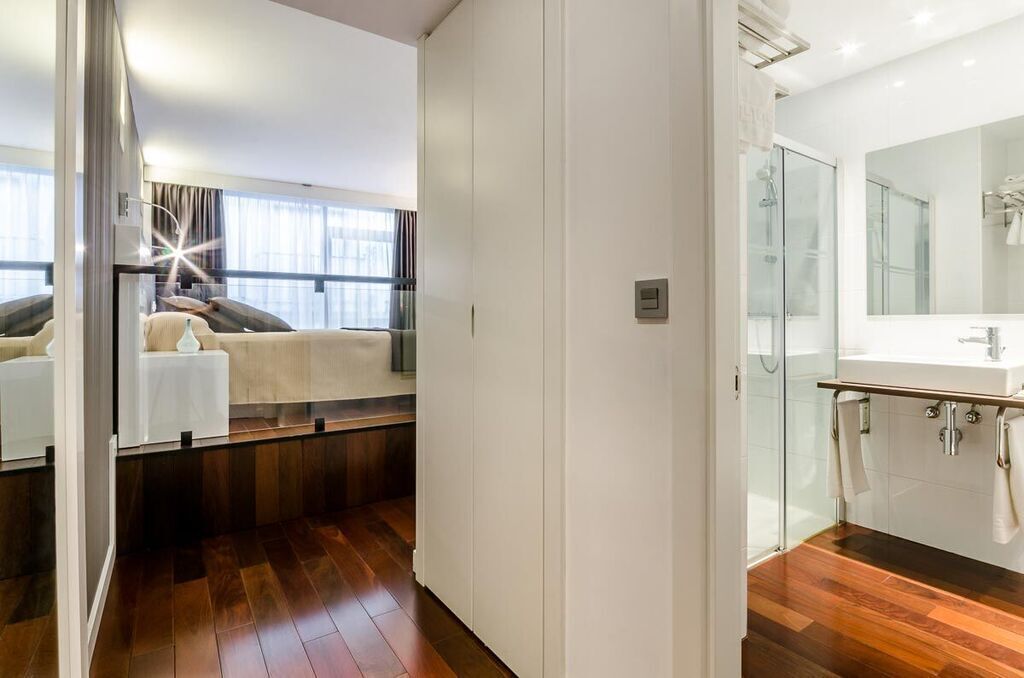 A modern hotel room with a large bed, a sitting area with a sofa and a coffee table, a separate bathroom with a glass shower, and a large window providing natural light.