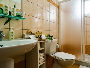 A bathroom with a sink, toilet, and shower. There are towels and toiletries on the shelves. - Tsambikos Apts (Rhodes)