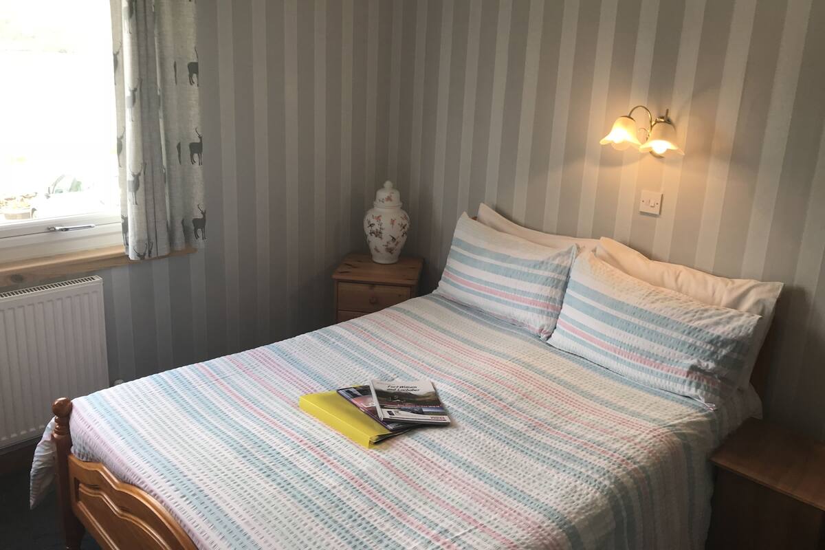 Un dormitorio con pared a rayas, una cama con ropa de cama a rayas, una mesita de noche con un jarrón y una ventana con cortinas.
