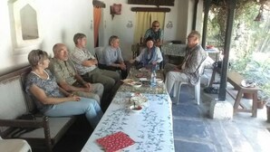 Un grupo de personas sentadas alrededor de una mesa con comida y bebidas, en una habitación con piso de baldosas y ventana con vista a áreas verdes.