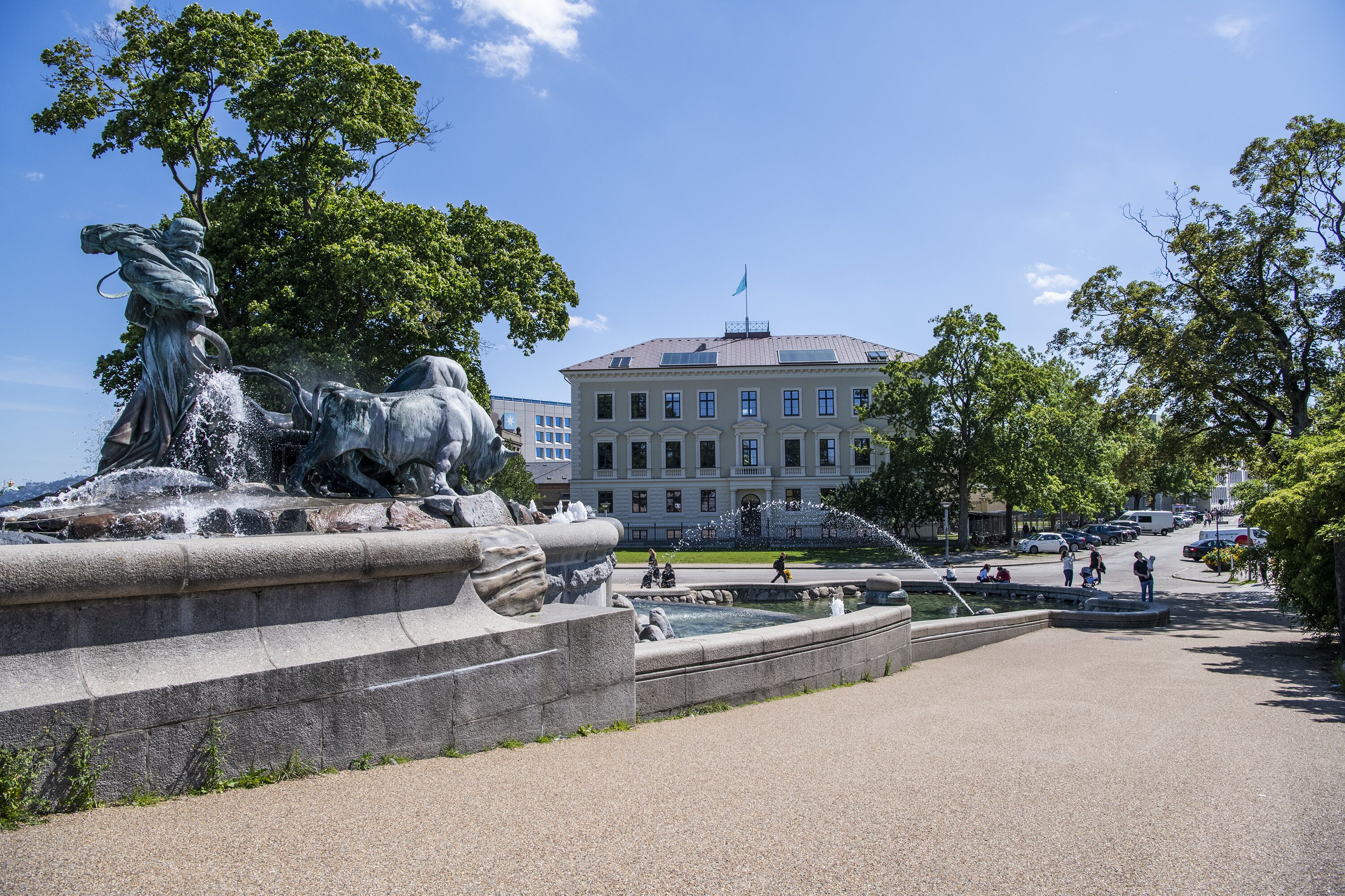 A public square with a fountain, a large building in the background, and a clear sky.