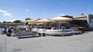 Een buitenzitgedeelte met banken en tafels, een overdeeld deel met parasols, en een gebouw met borden op de achtergrond.