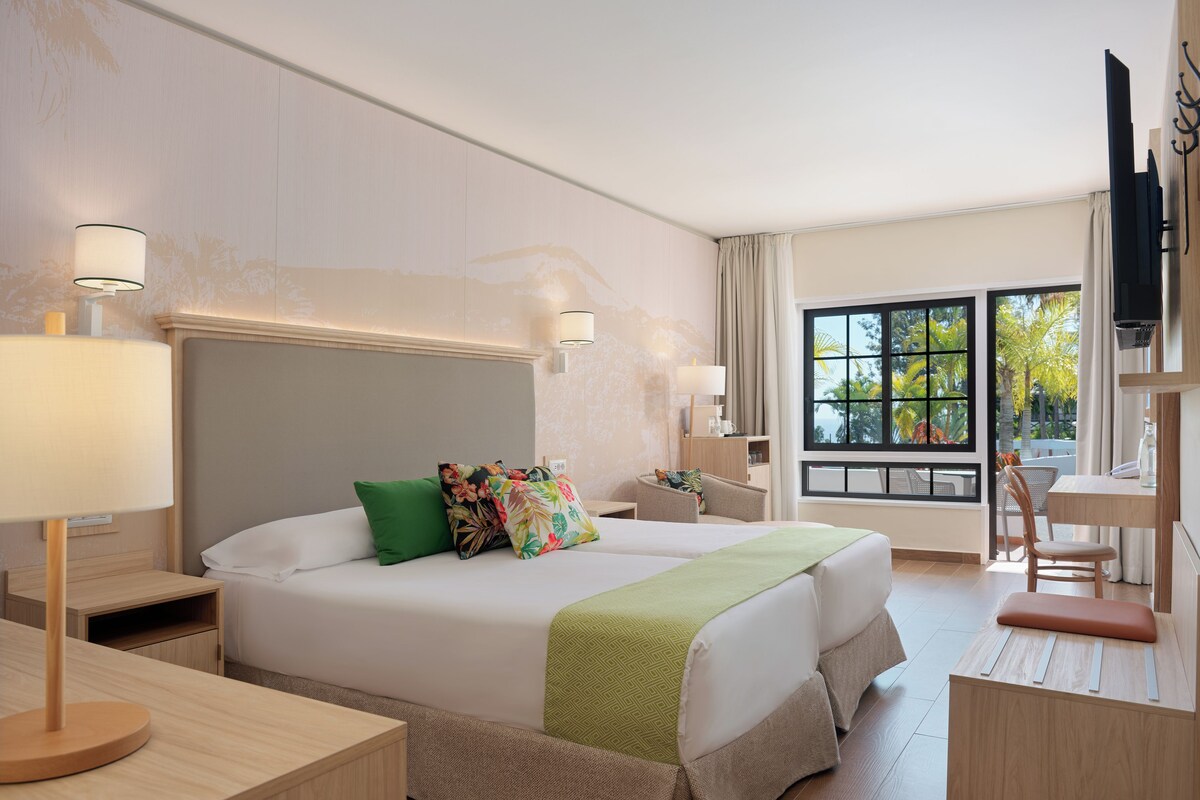 A hotel room with a large bed, a desk, a chair, a TV, and a window with a view of palm trees.