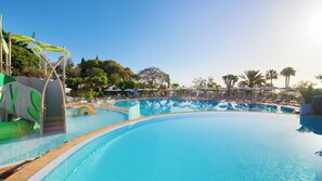 Una piscina grande con tobogán, rodeada de sillas de descanso y sombrillas, con árboles al fondo.