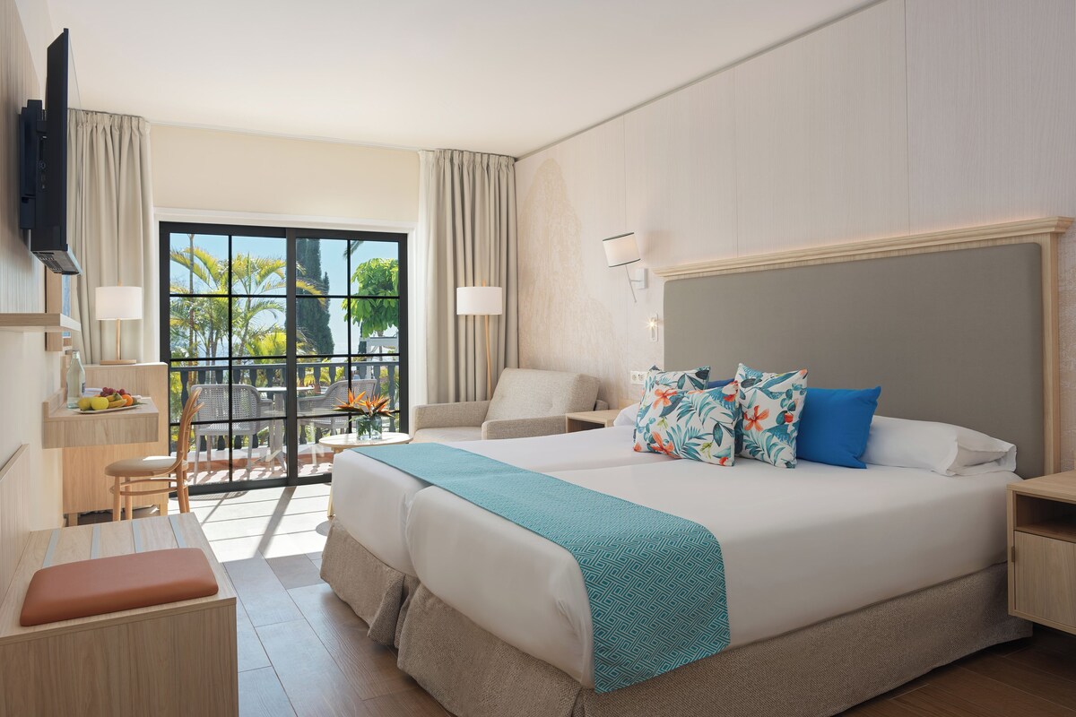 A hotel room with a large bed, a desk, a chair, a TV, and a balcony with a view of palm trees.